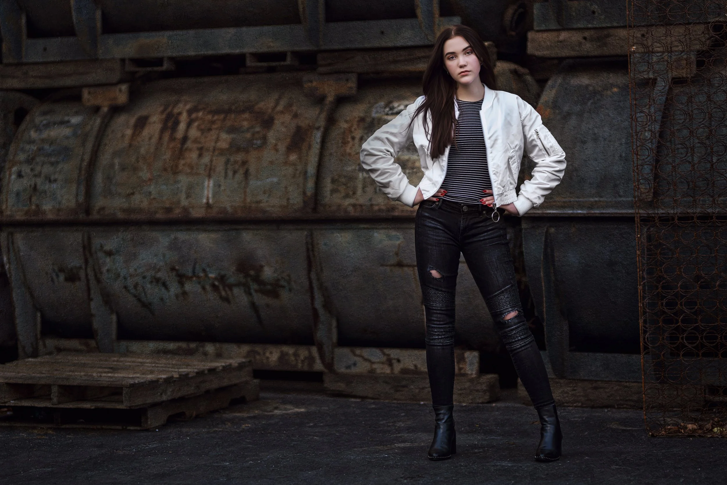 A young woman with long dark hair stands with her hands on her hips in front of rusty industrial equipment, wearing a white bomber jacket, black ripped jeans, and black ankle boots.