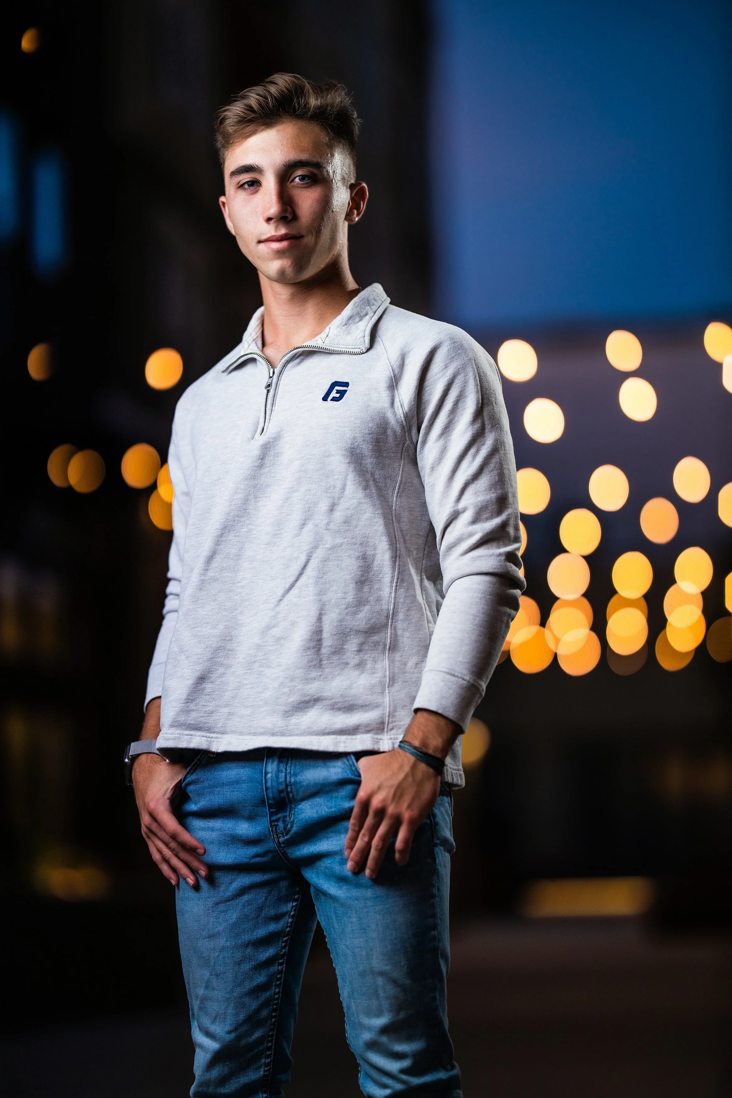 A young man in a light gray quarter-zip sweatshirt and jeans standing outdoors at night with blurred yellow string lights in the background.