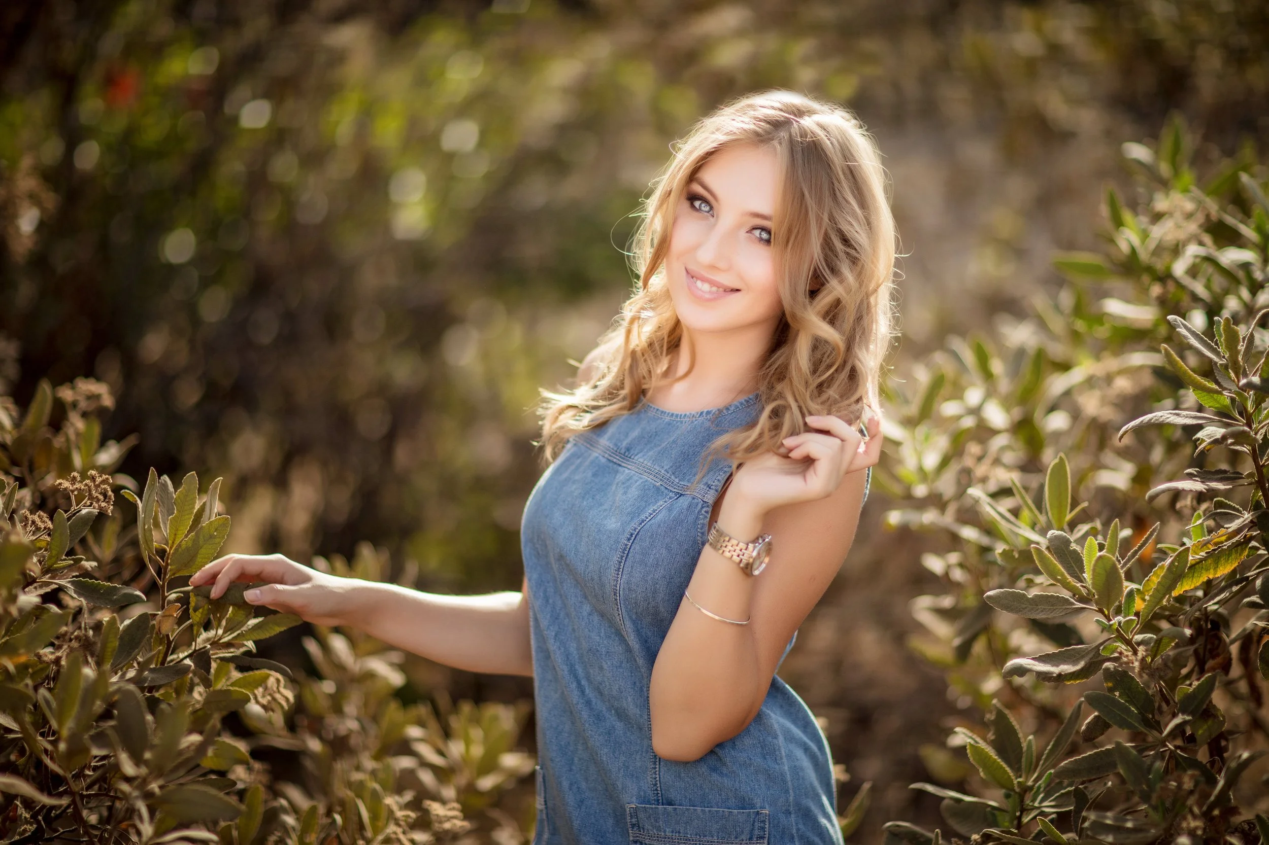 A young woman with wavy blonde hair and blue eyes smiling at the camera, wearing a sleeveless denim dress, in an outdoor setting surrounded by greenery with sunlight filtering through the leaves.