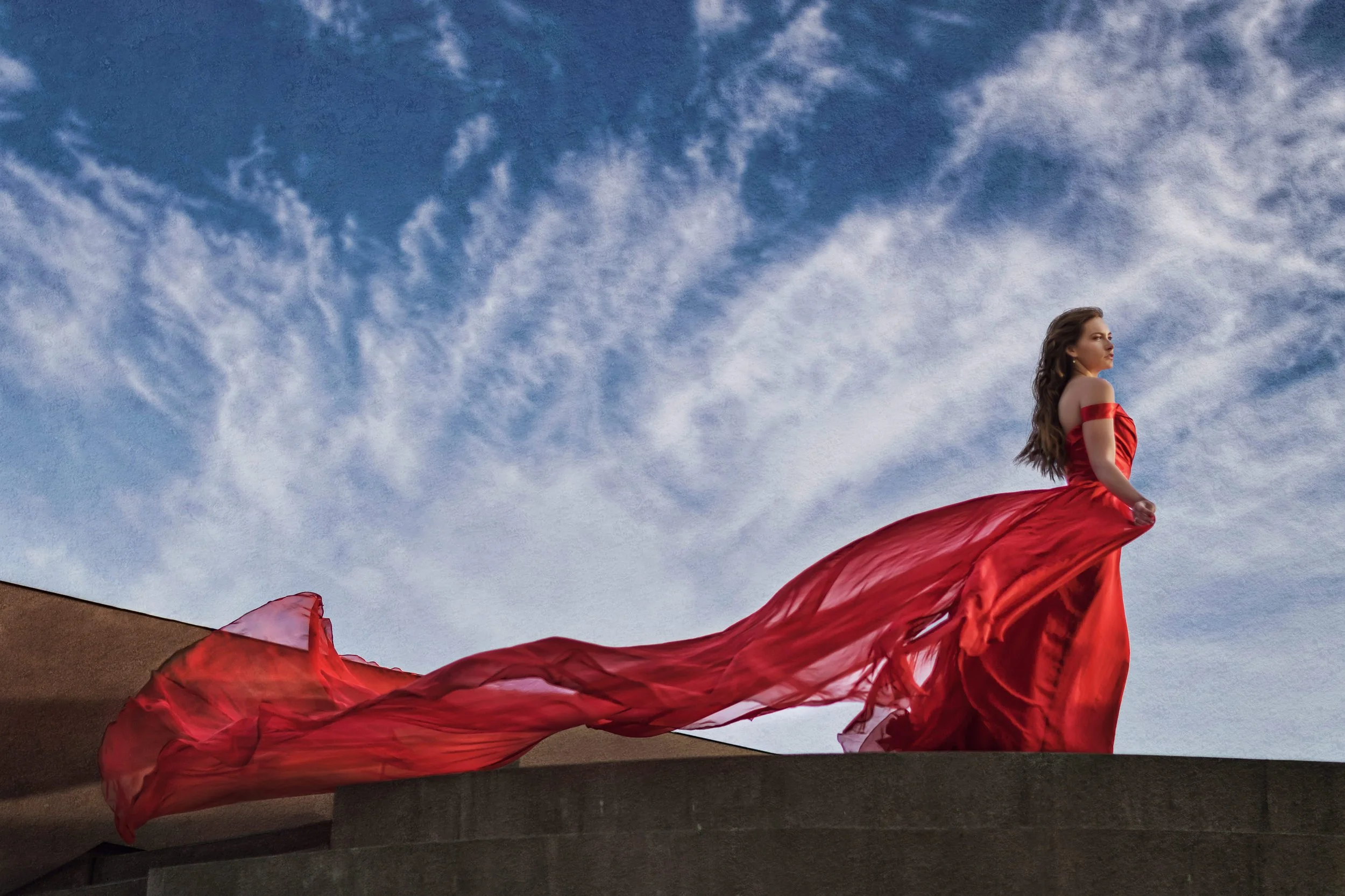 A woman in a flowing red dress standing on a ledge against a cloudy sky.