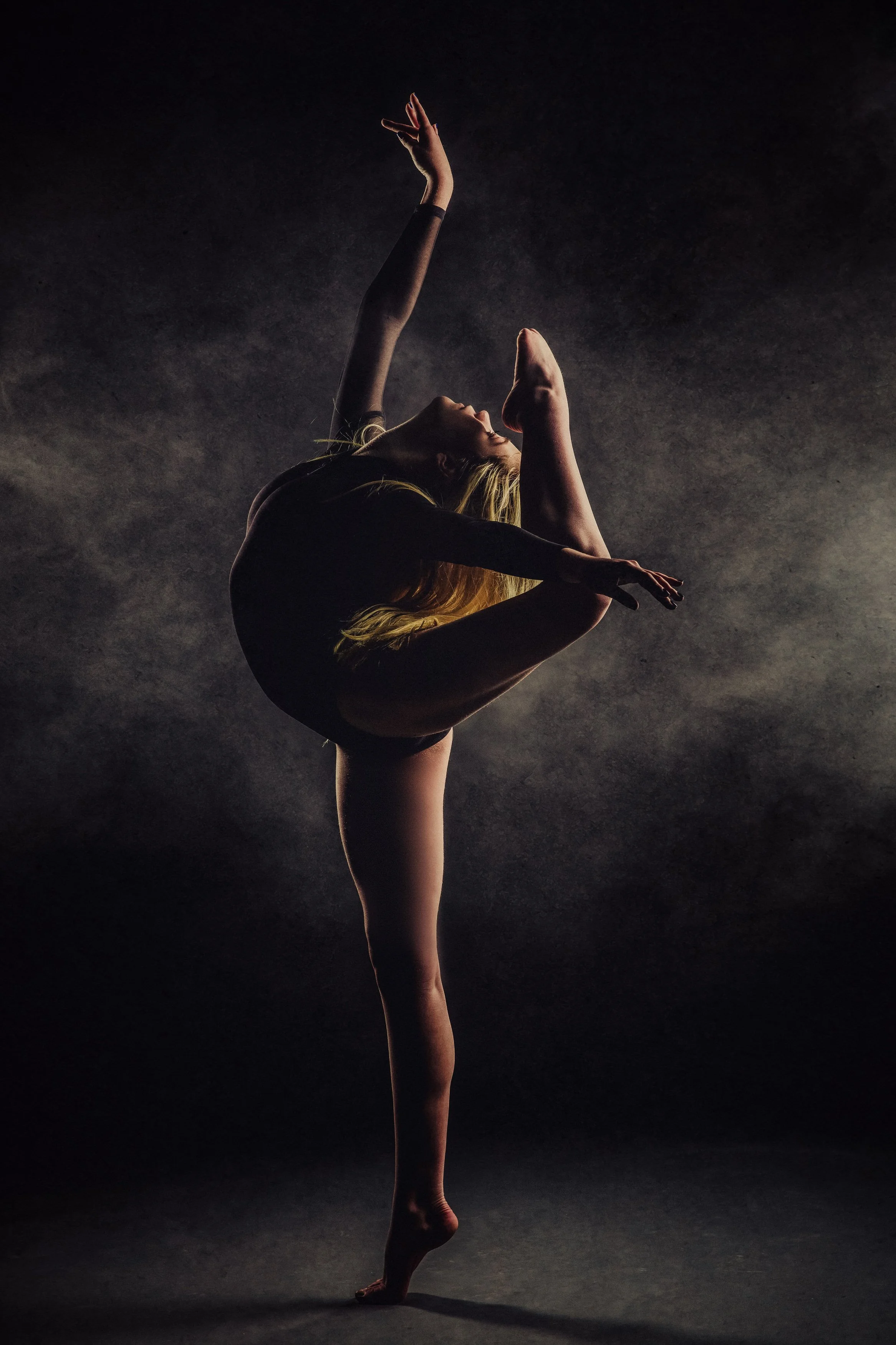 A female dancer performing a balancing pose in a dark studio, with one leg raised and bent, and her arm extended upward.