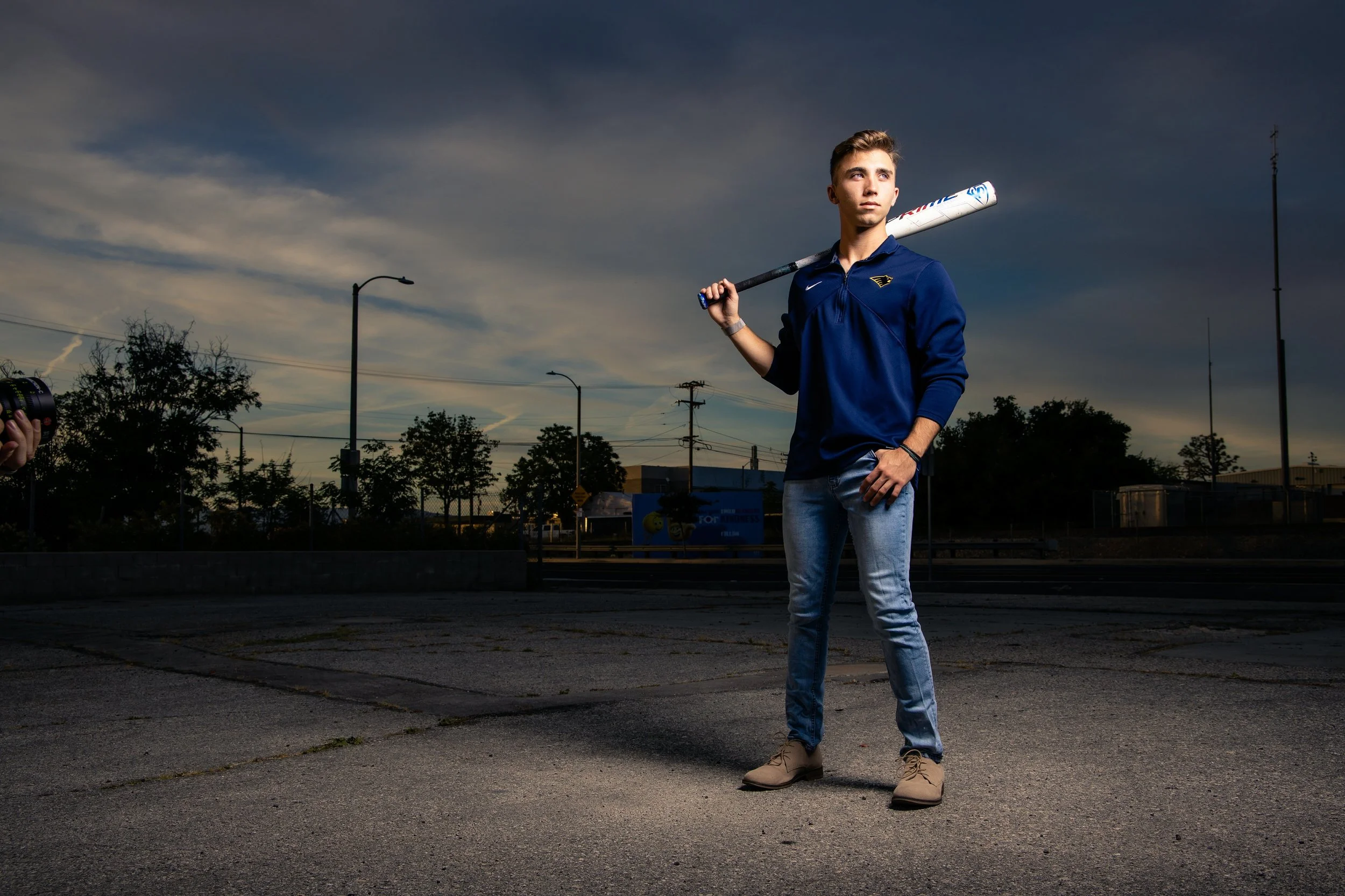 Young man in a navy hoodie and jeans standing outdoors during dusk, holding a baseball bat resting on his shoulder, with a serious expression, streetlights, power lines, and trees in the background.