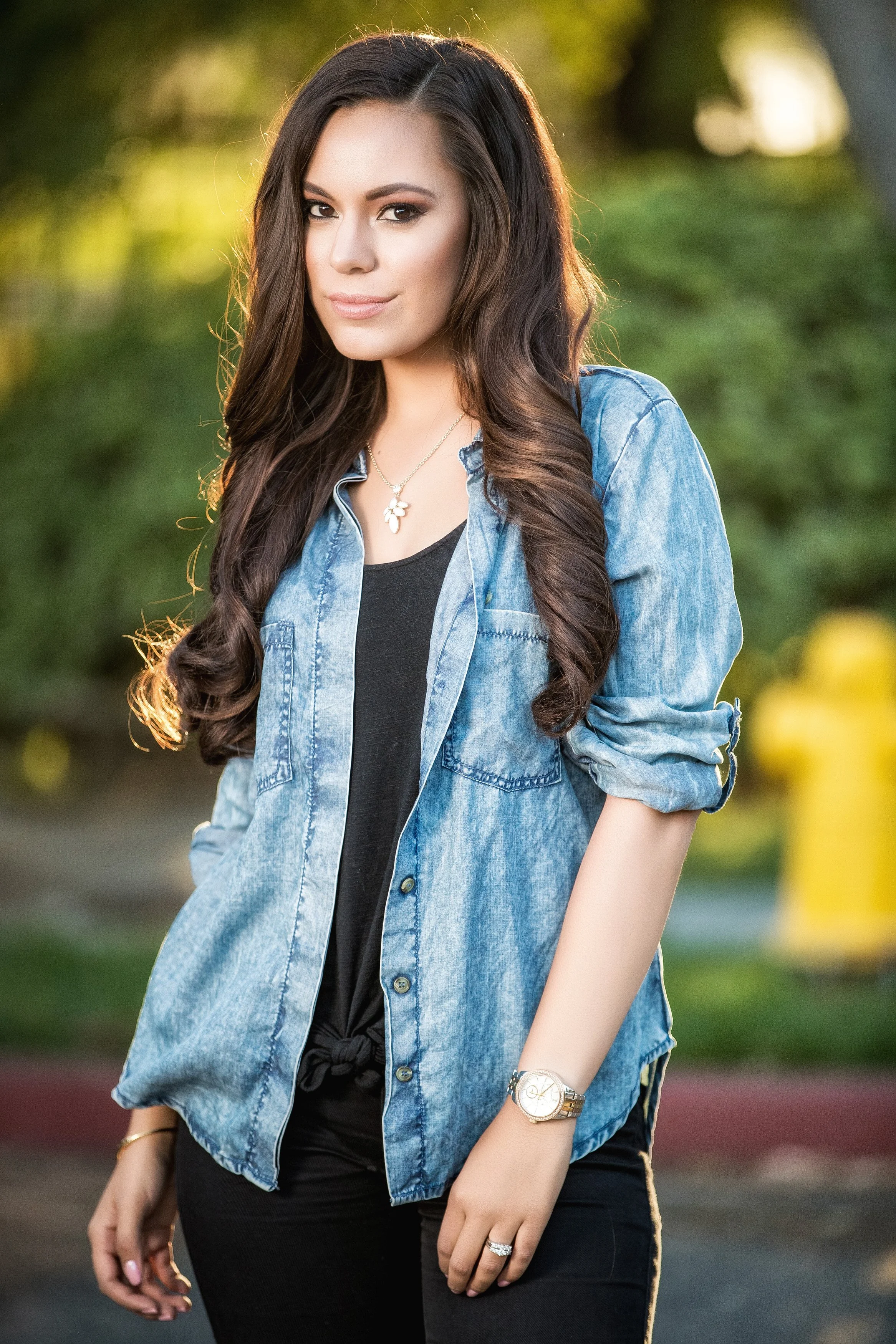 A young woman with long, wavy dark hair, wearing a denim shirt over a black top, standing outdoors during golden hour with greenery in the background.