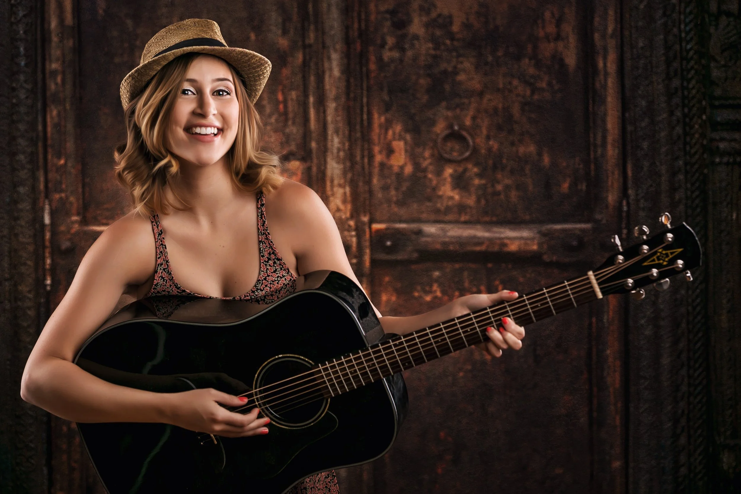 A young woman with shoulder-length blonde hair wearing a straw hat, a sleeveless floral dress, smiling and holding a black acoustic guitar, standing in front of a wooden background.