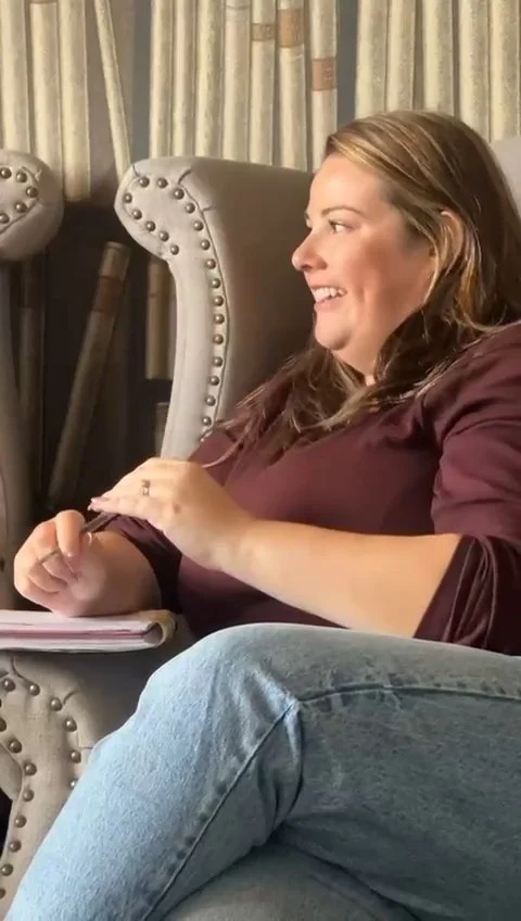 A woman sitting in a comfortable chair with a smile, looking to her right, with book shelf printed wallpaper in the background.