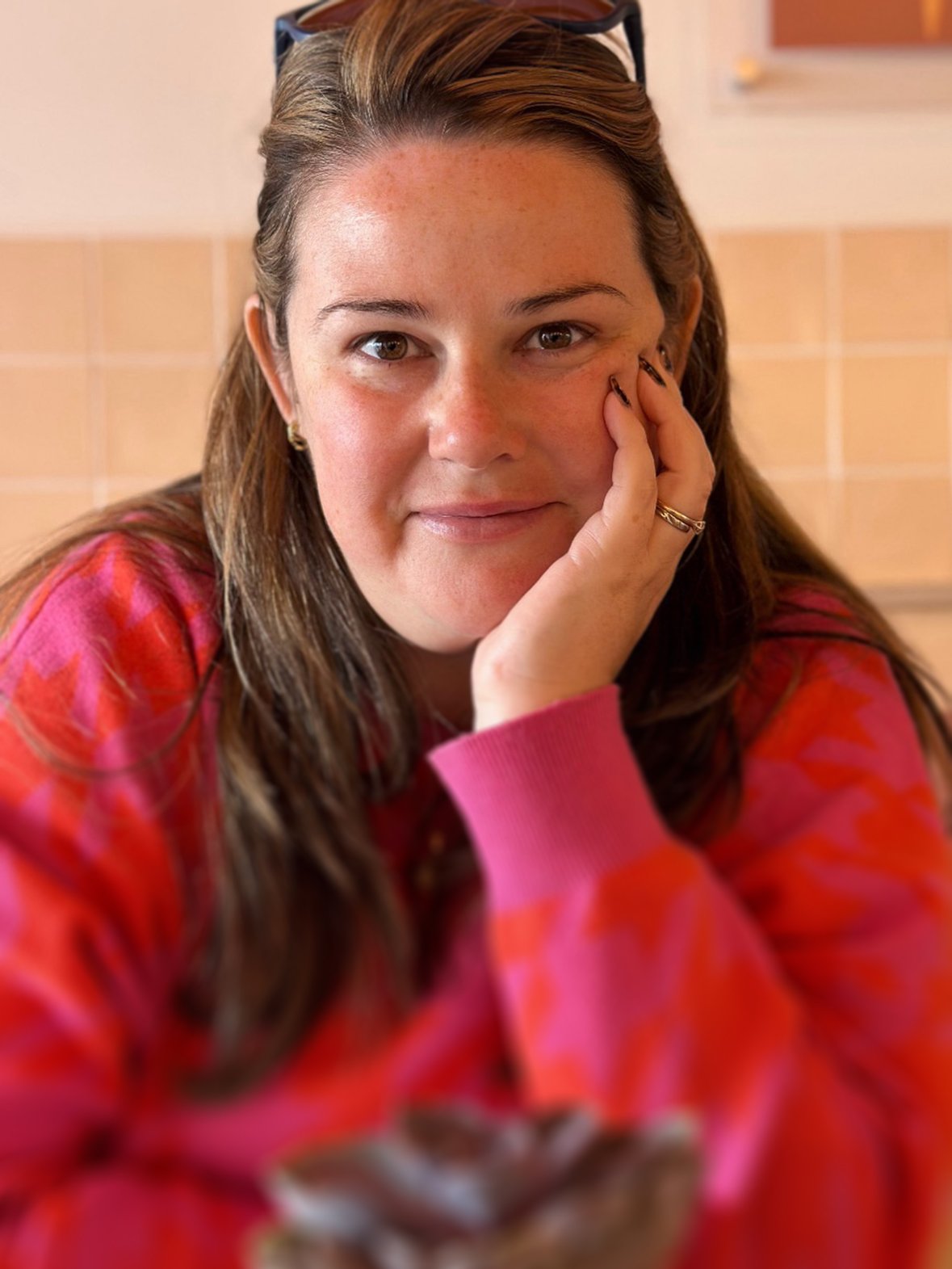 A woman with brown hair, wearing a pink and red patterned top, is resting her face on her hand and looking at the camera, with a slight smile.