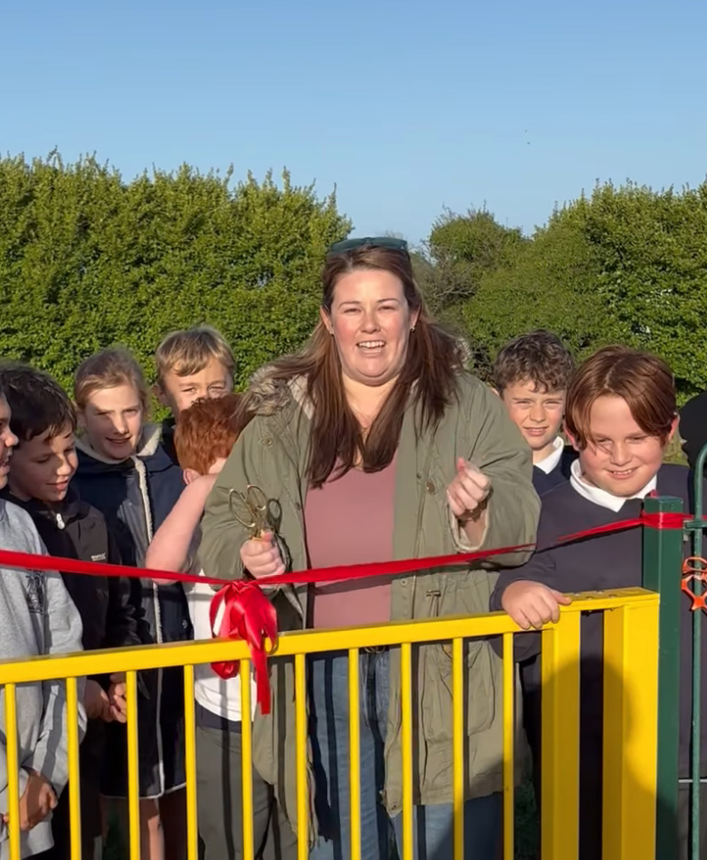 Woman cutting a red ribbon with scissors at a ribbon-cutting ceremony at a new park, surrounded by children, outdoors on a sunny day.