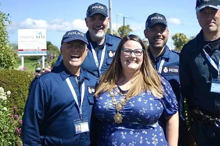 Group of five people outdoors, four men in police uniforms and one woman in a blue floral dress with glasses, all smiling.