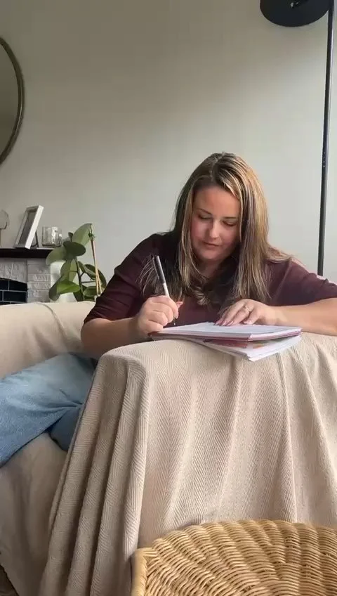 A woman sitting on a couch, working on papers on a small table covered with a beige cloth, with a plant and picture frame in the background.