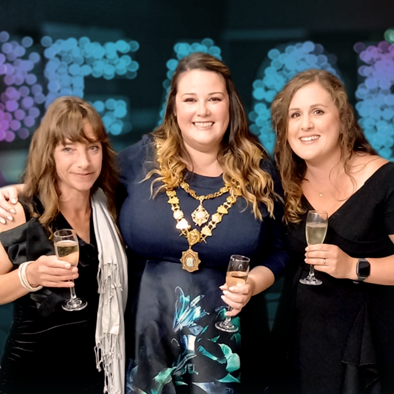 Three women at a celebration, holding glasses of champagne, with colorful bokeh lights in the background.