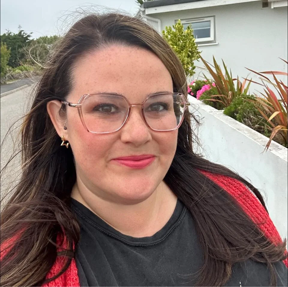 A woman with long dark hair wearing glasses, earrings, a black shirt, and a red cardigan, standing outdoors near a white building with plants and flowers.