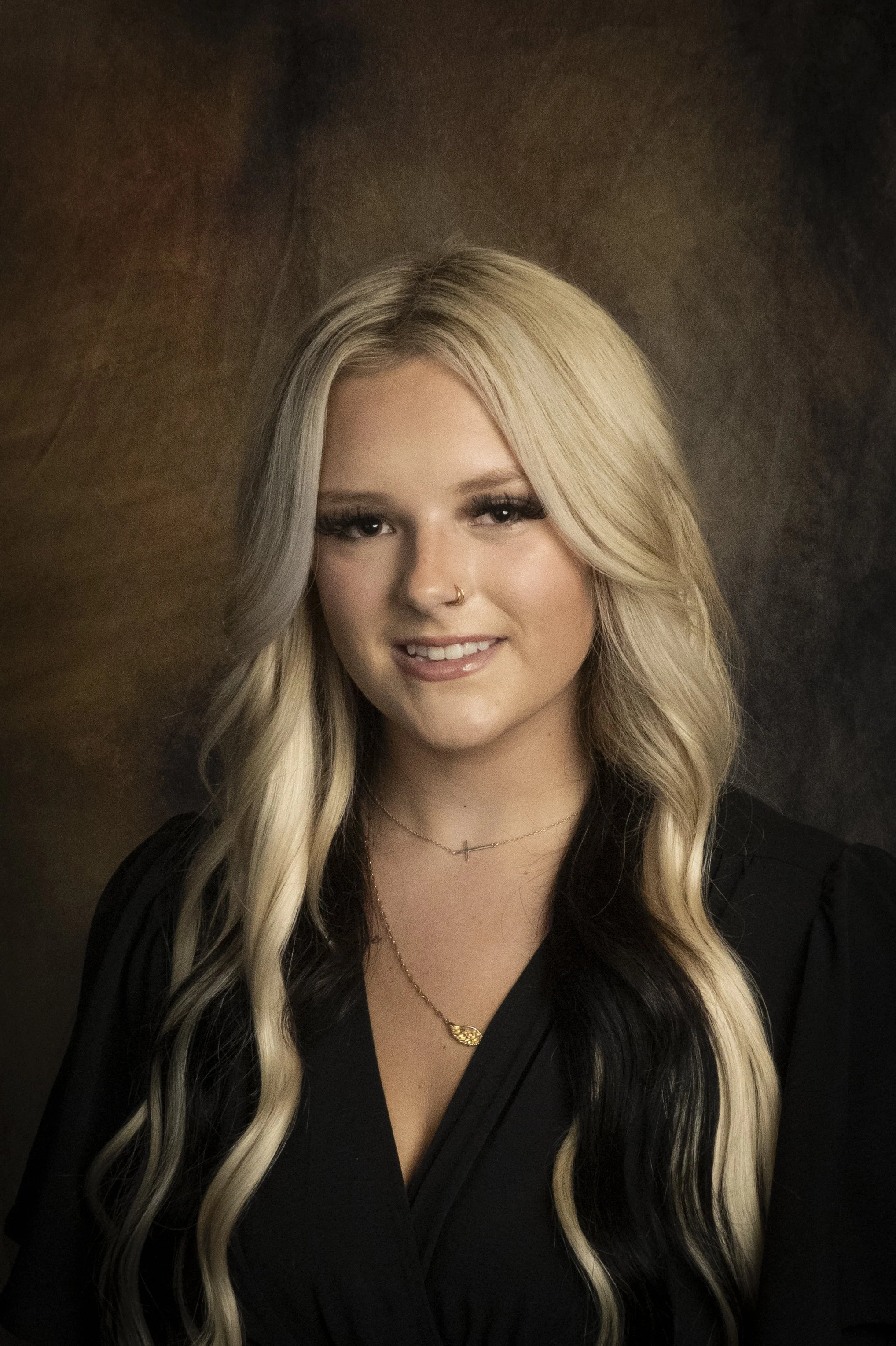Portrait of a young woman with long wavy blonde and black hair, wearing a black top, gold and silver necklaces, and a nose ring, smiling softly against a dark brown background.