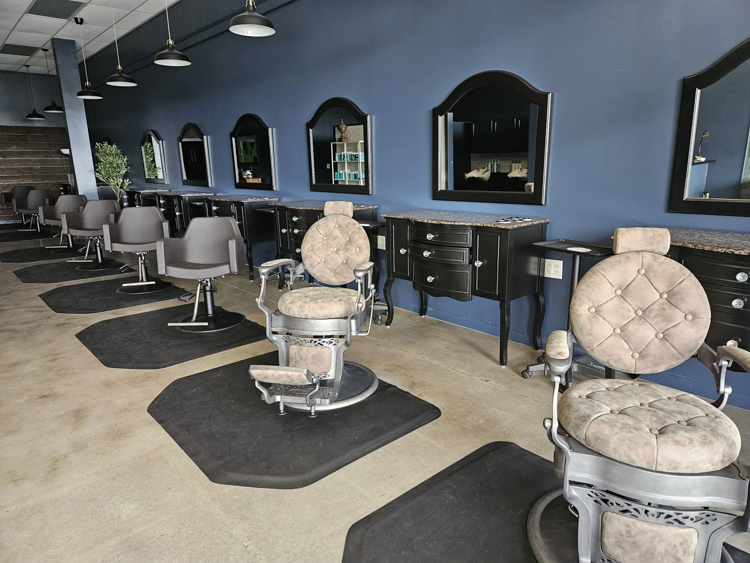 Empty salon chairs in front of black styling stations with mirrors on a blue wall.