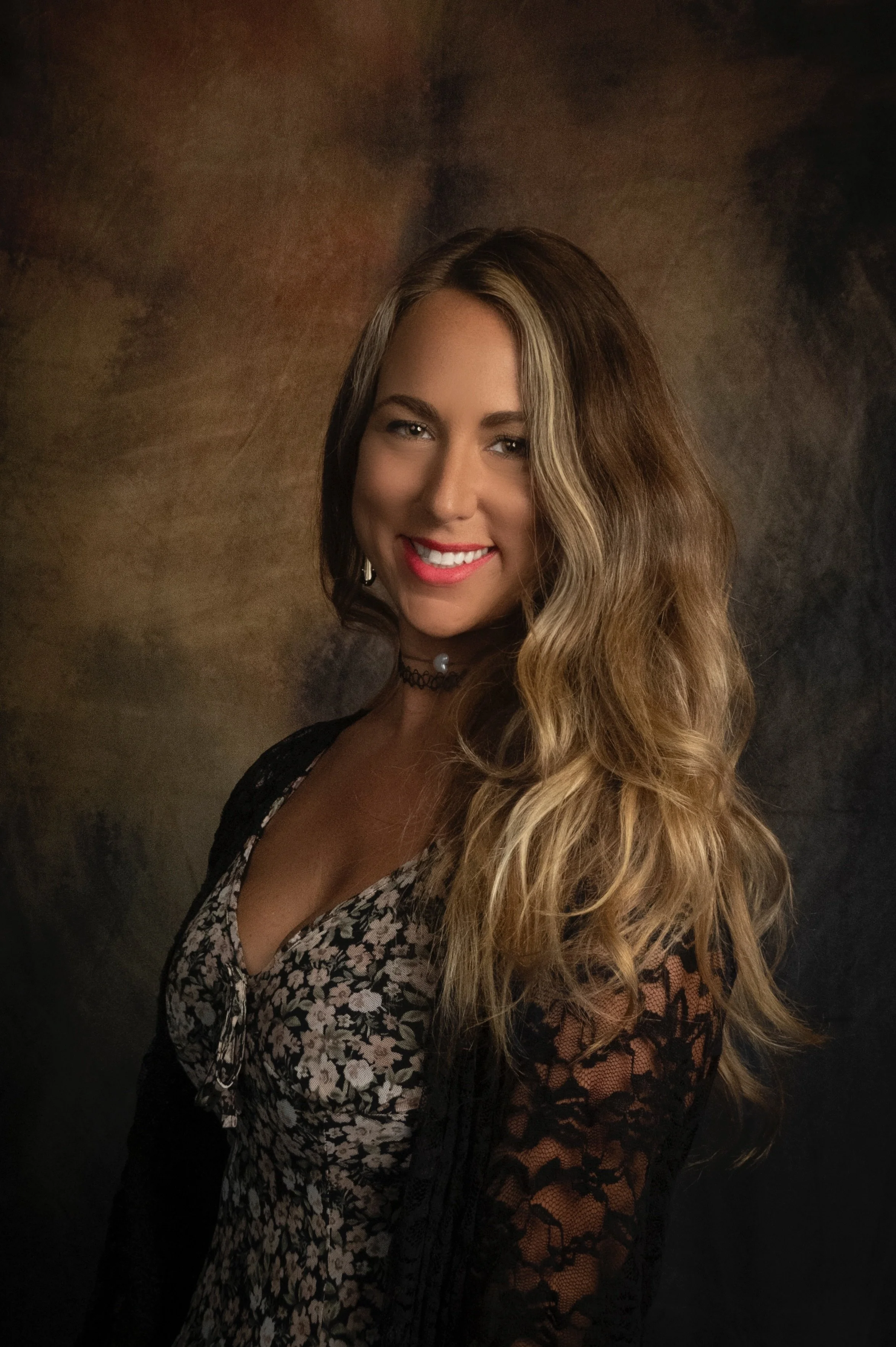 A young woman with long, wavy blonde hair smiling and looking at the camera, wearing a black floral dress with lace sleeves and a choker necklace, against a dark, textured background.
