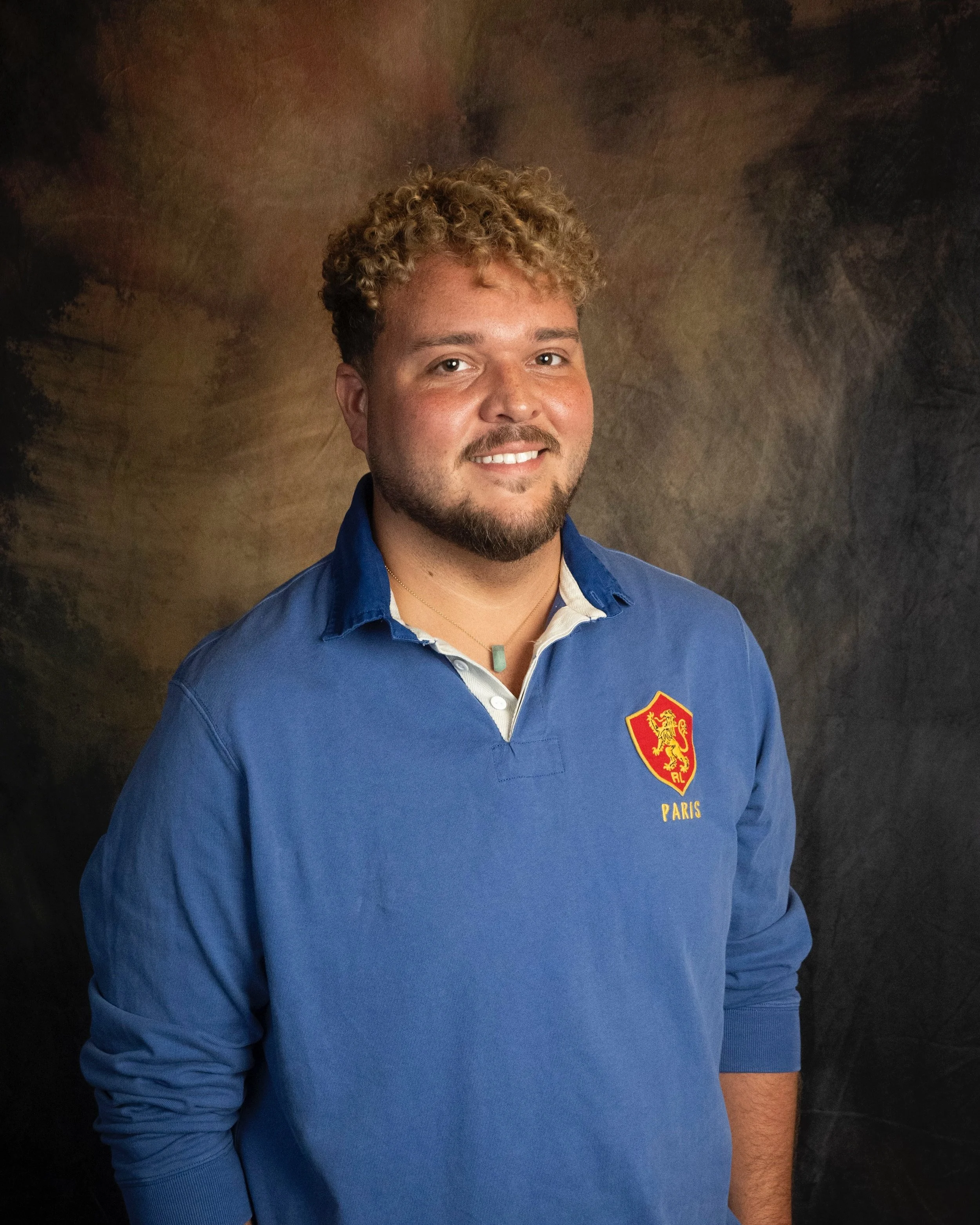 A man with curly blonde hair and a beard smiling, wearing a blue rugby shirt with a crest and the word 'Paris' on it, standing against a dark, textured background.