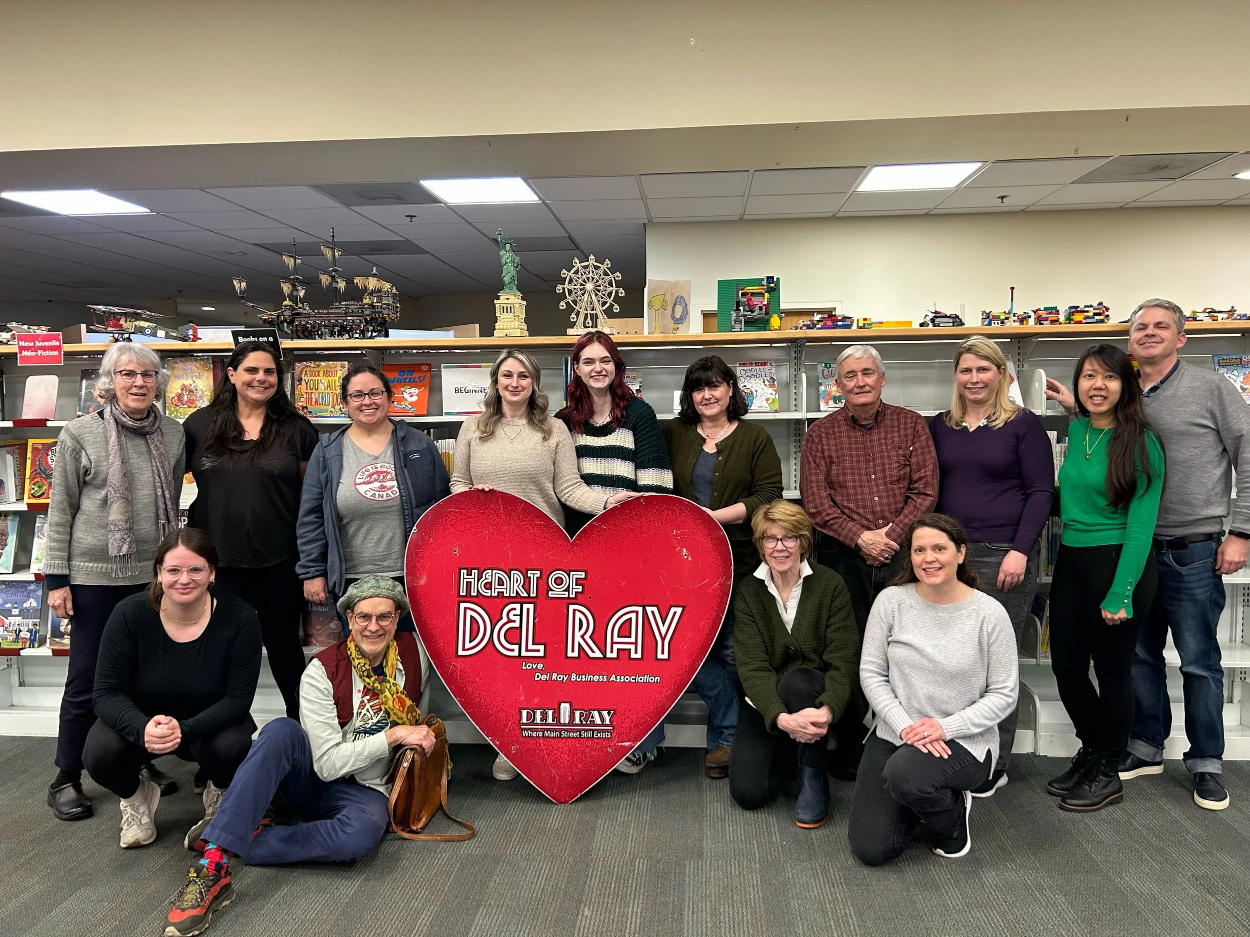 Group of fifteen people, diverse in age and ethnicity, posing in a library with bookshelves behind them. They are smiling and holding a large red heart-shaped sign that reads 'Heart of Del Ray' with additional text. Some individuals are kneeling, whi