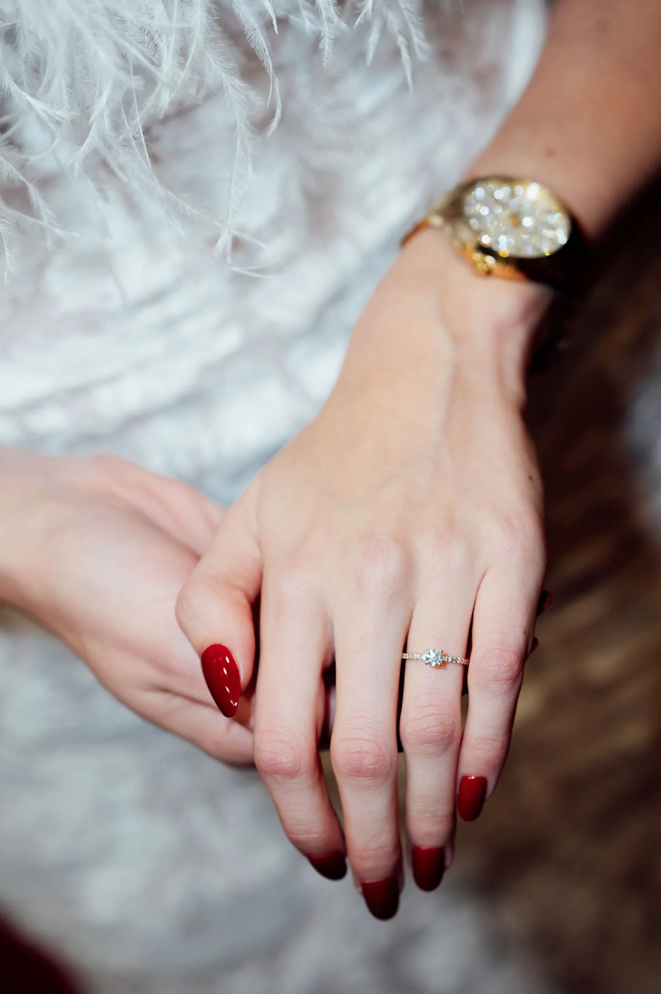 Nahaufnahme einer Hand mit einem Verlobungsring, rotem Nagellack und einem eleganten Handgelenk mit einer goldfarbenen Uhr, im Hintergrund weiße Federdeko.