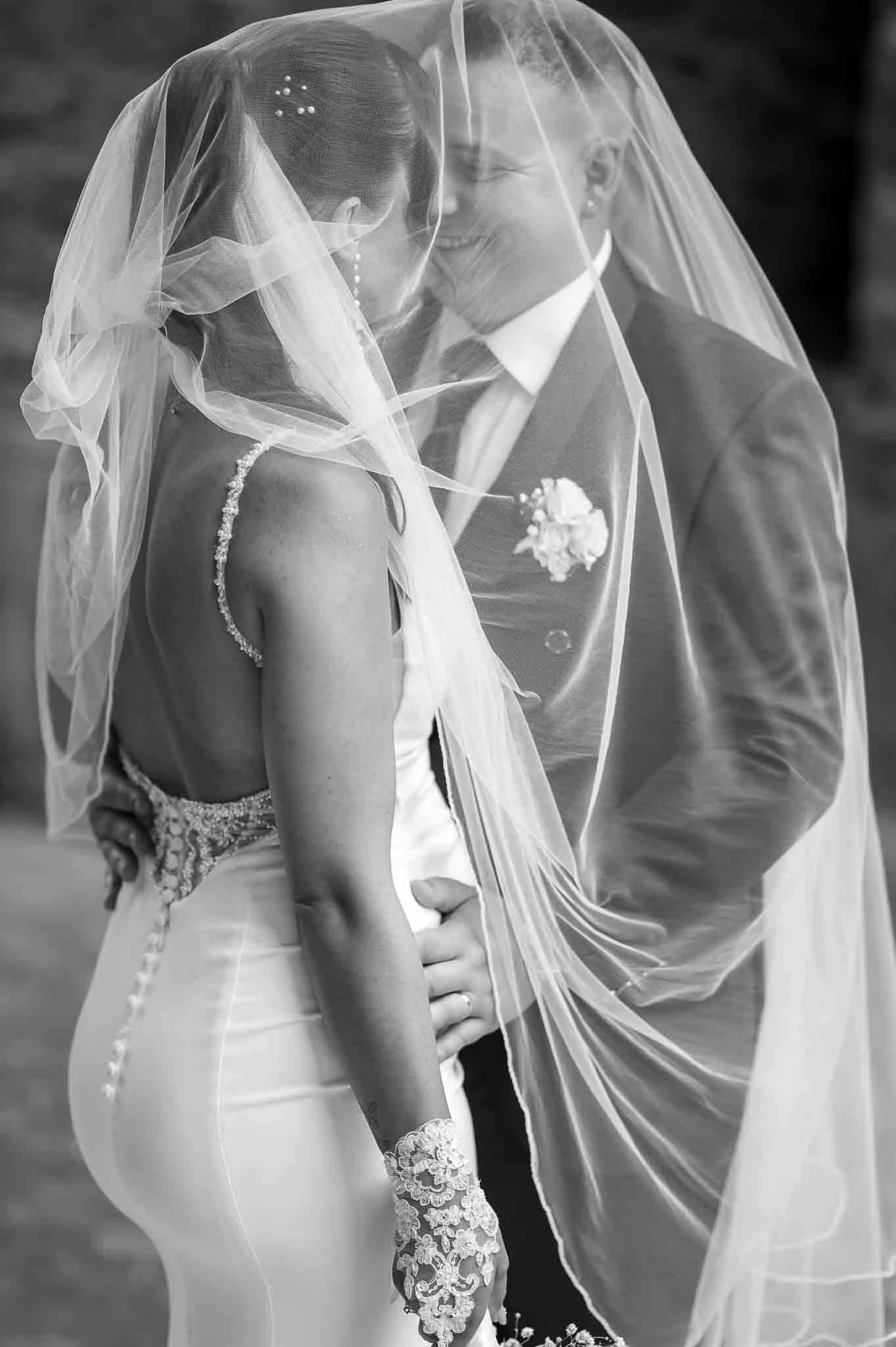 Braut und Bräutigam stehen sich nahe unter einem Schleier, die Braut trägt ein elegantes Kleid mit Spaghettiträgern und Handspitze-Accessoire, während der Bräutigam einen Anzug mit Fliege trägt, beide lächeln bei ihrer Hochzeitszeremonie.