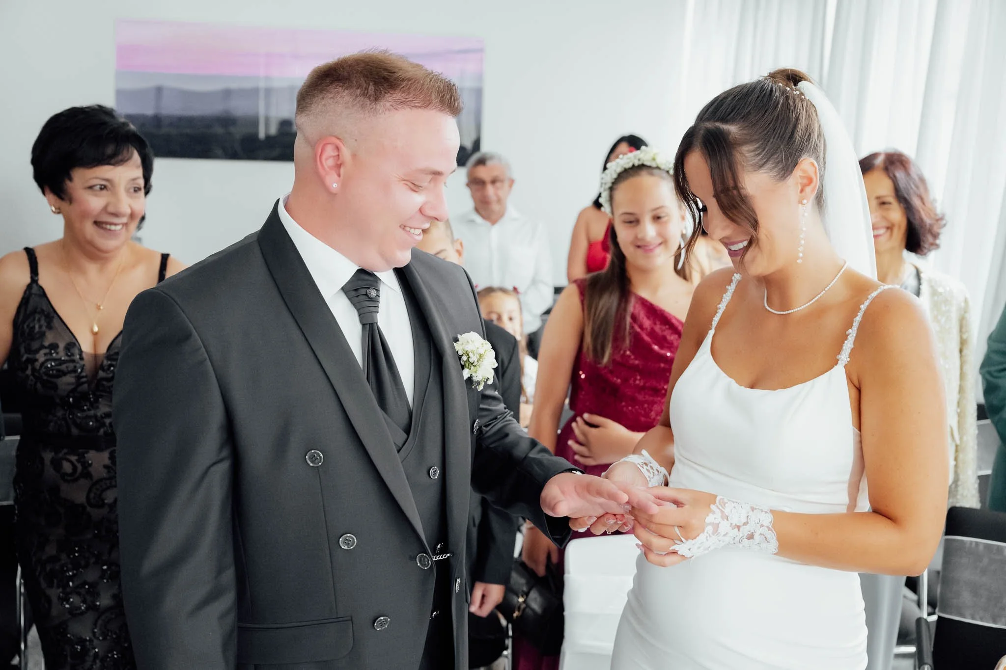 Braut und Bräutigam tauschen Eheringe bei Hochzeit, umgeben von lachenden Gästen.