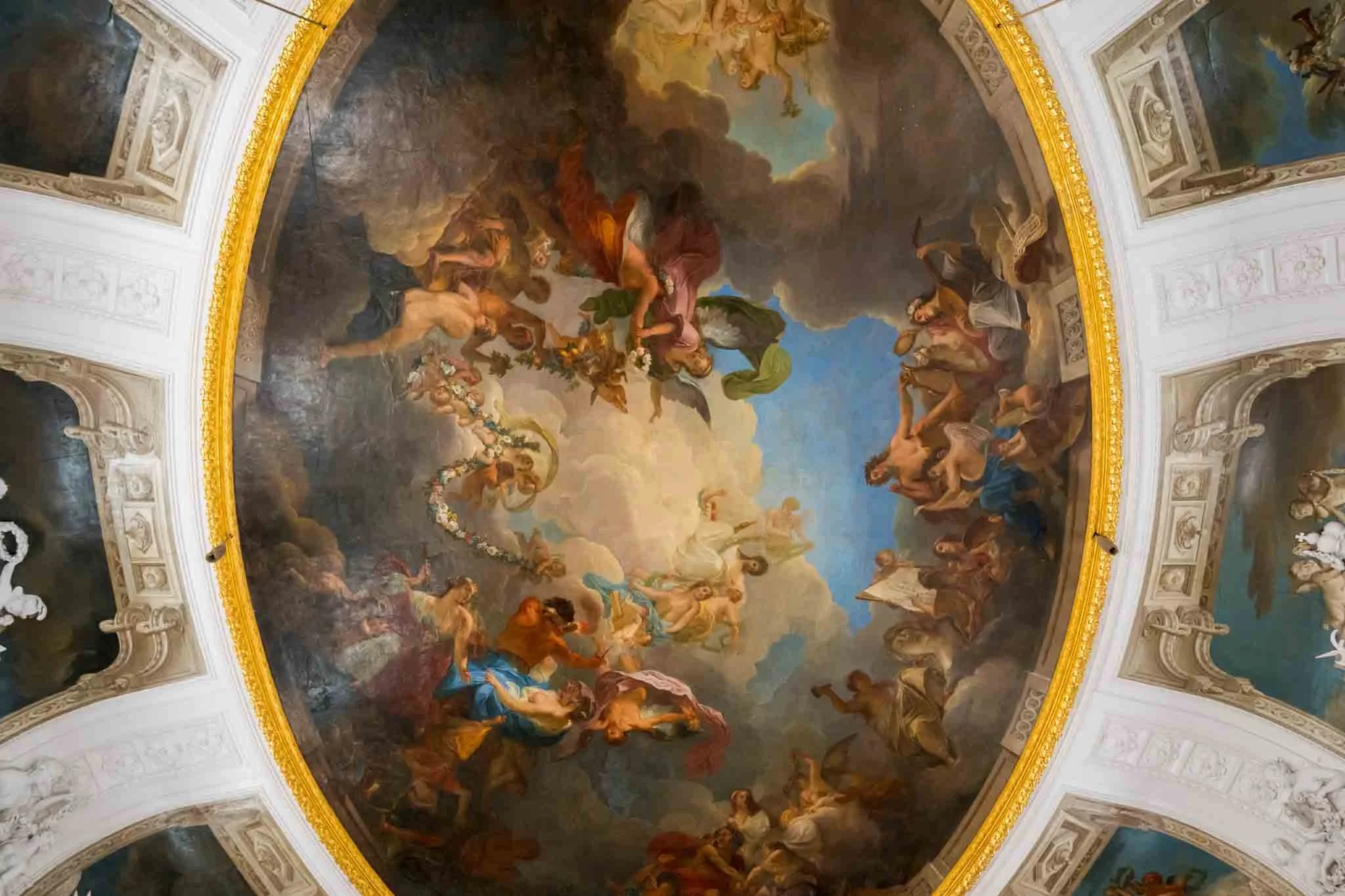 Ein aufwendiges dekoloriertes Barock-Wandgemälde auf einer gewölbten Decke, das Szenen mit mythologischen oder religiösen Figuren in dramatischem Moment zeigt, umrahmt von goldfarbenem Rahmen und weißen, dekorativen Elementen.