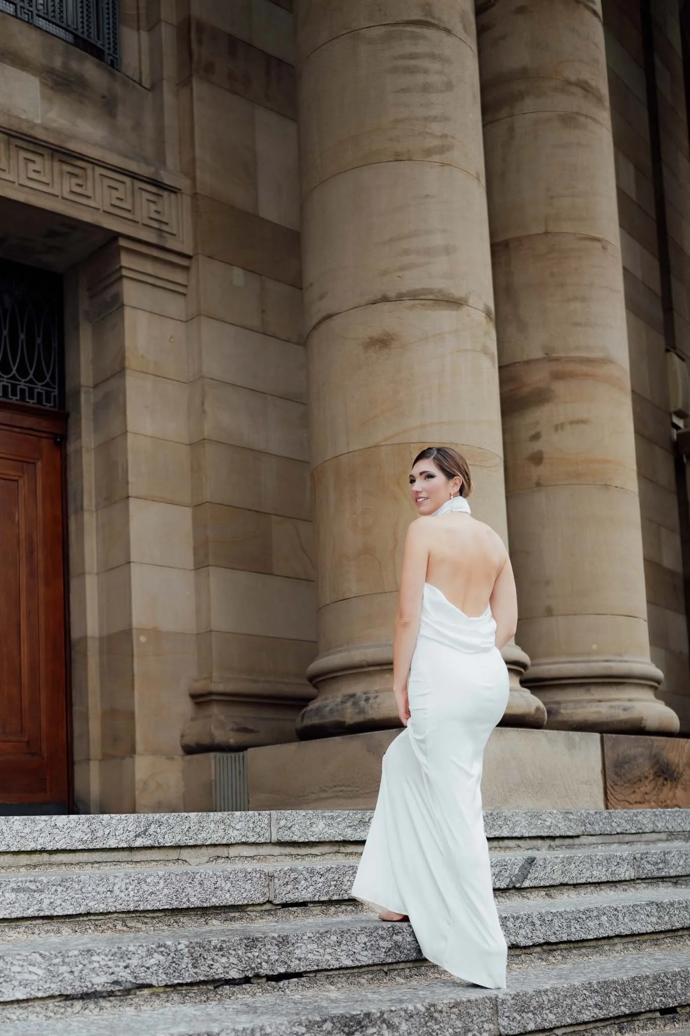 Eine Frau in einem weißen Abendkleid steht auf Steinstufen vor einem antiken Gebäude mit großen Säulen.