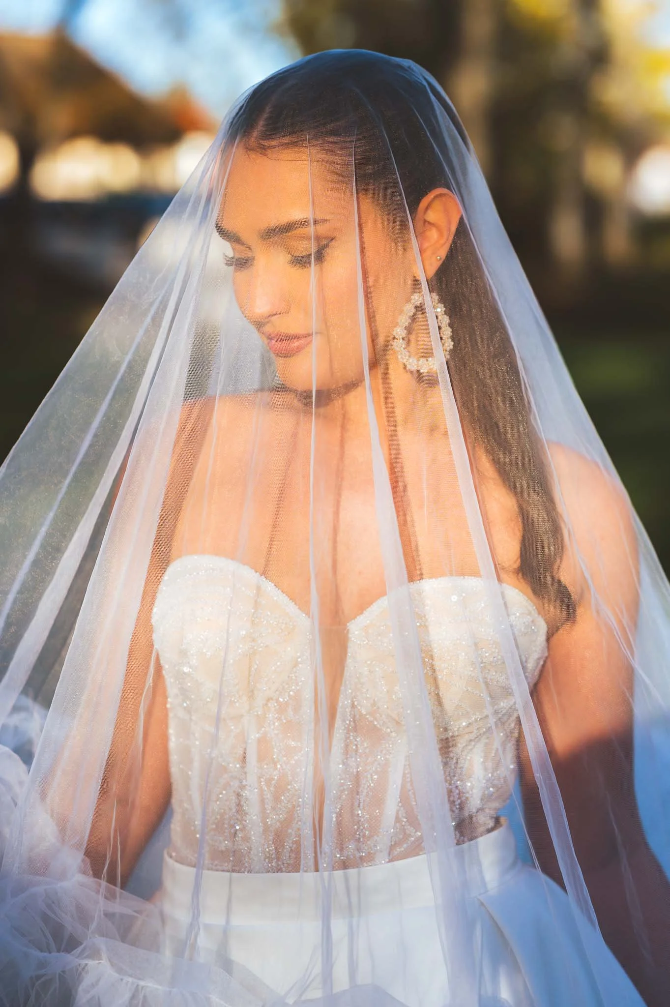 Braut mit Schleier und Ohrringen, trägt ein weißes Brautkleid mit Pailletten, im Freien bei Sonnenuntergang, nachdenklich.