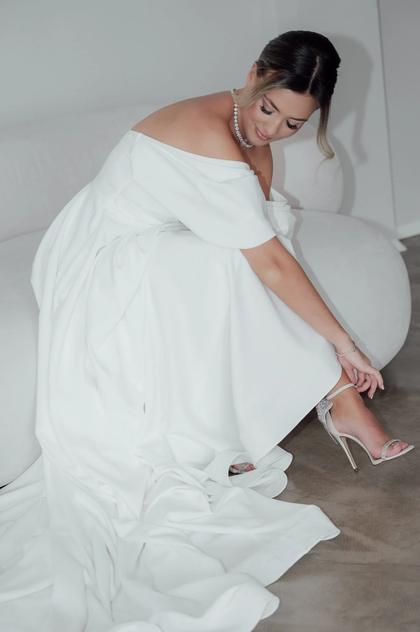 Frau in einem weißen Kleid, sitzt auf einem Sofa und zieht ihren Schuh aus.