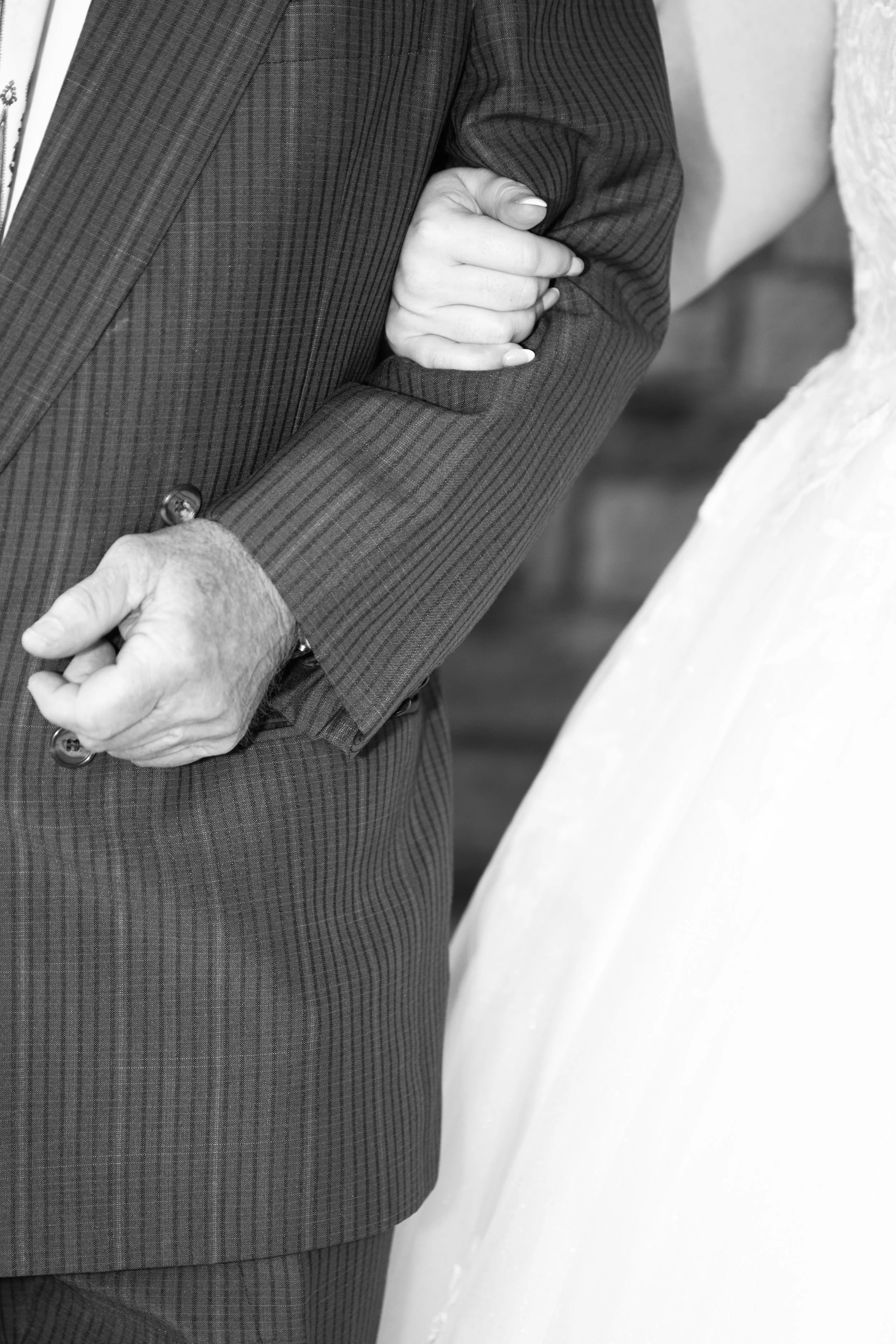 Close-up of a person's upper body, wearing a dark striped suit, with hands clasped in front, and a second person in a white dress standing nearby.