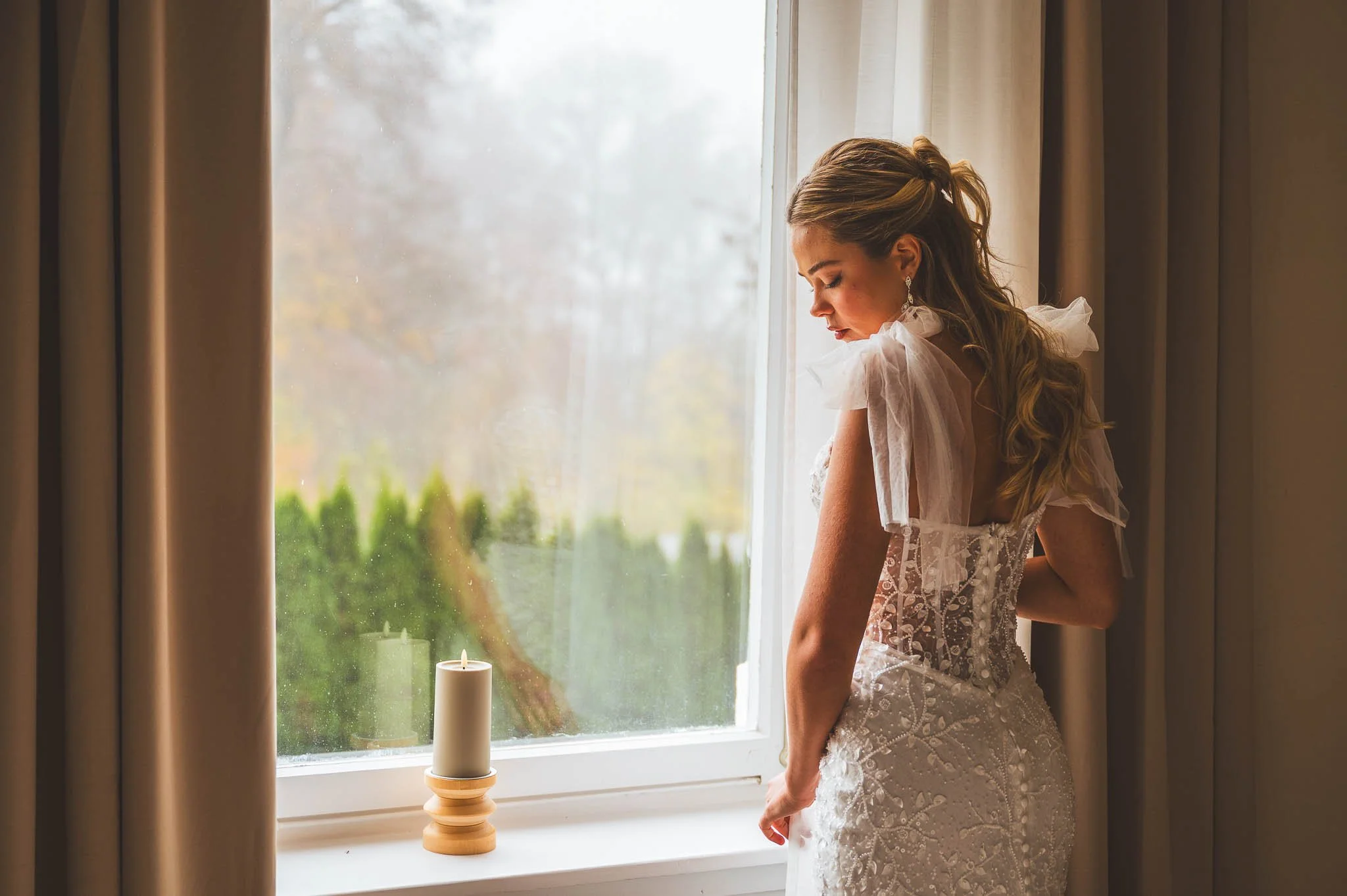Junge Frau im Brautkleid steht an einem Fenster mit Kerze, schaut nach draußen.