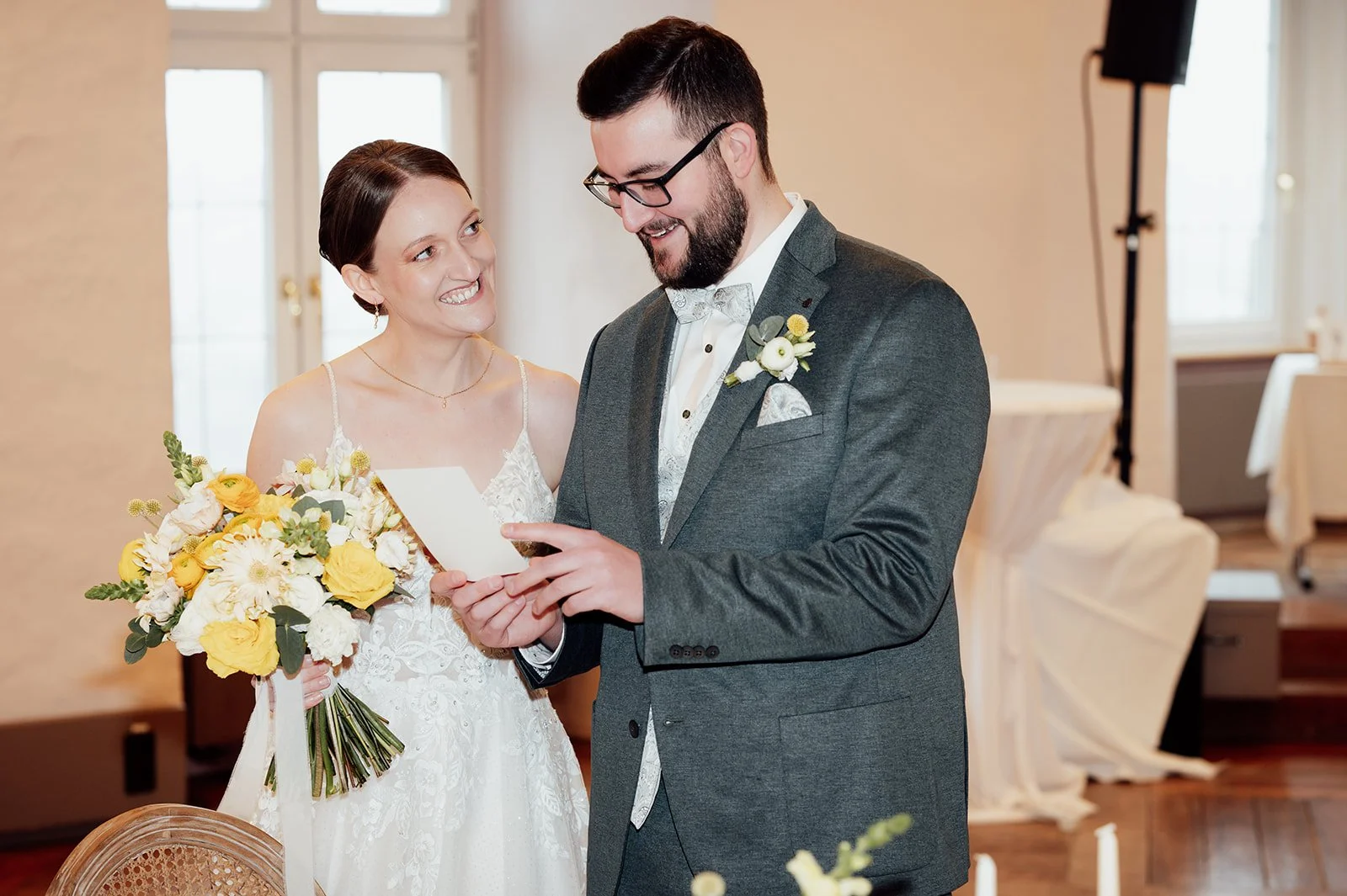 Eine Braut blickt einen Mann im Anzug lachend an, der gerade eine Karte liest und dabei selbst lacht.