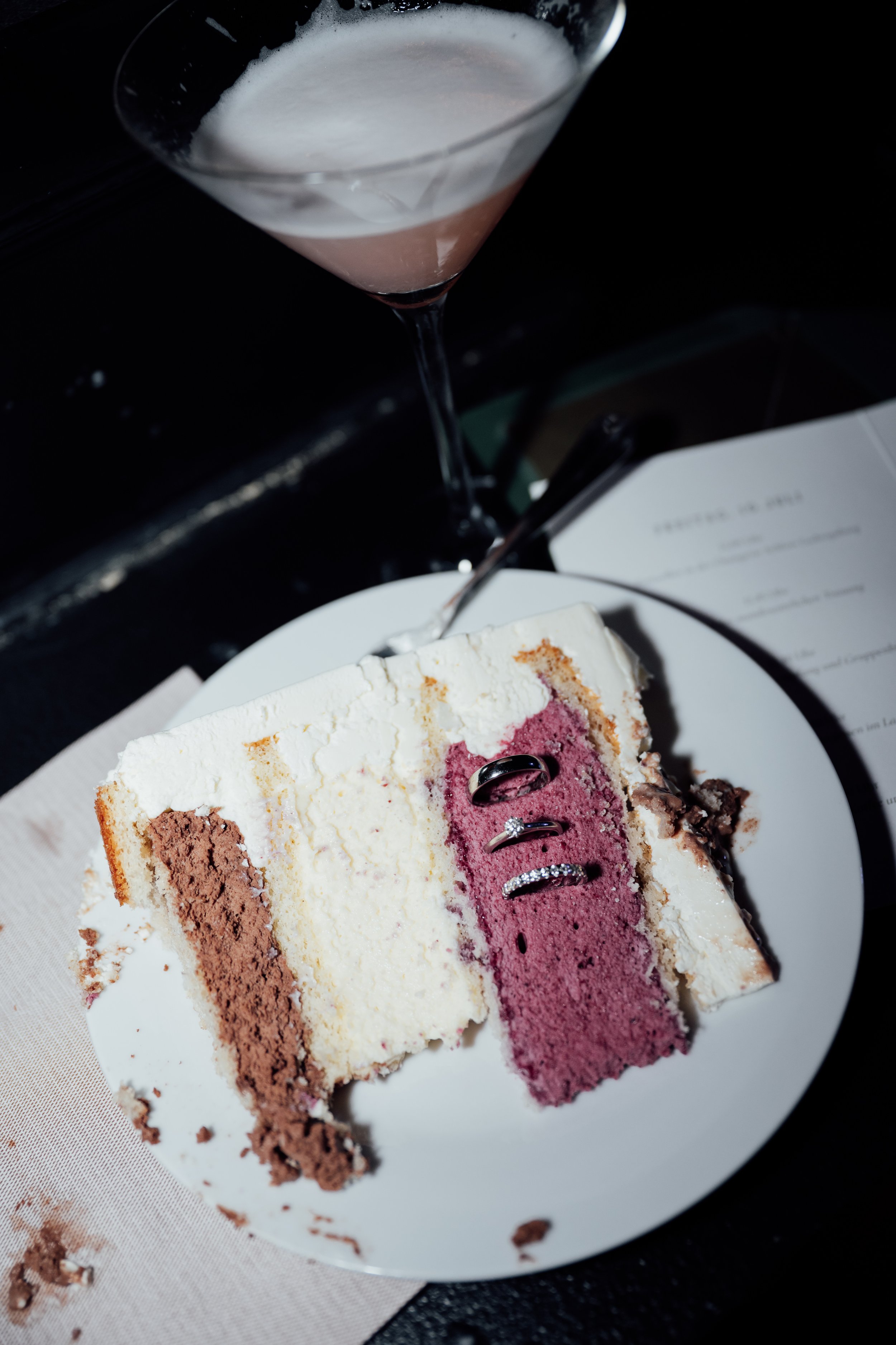 A slice of layered cake with white, pink, and chocolate layers, topped with utensils and wedding rings, on a white plate with a drink in a cocktail glass nearby.