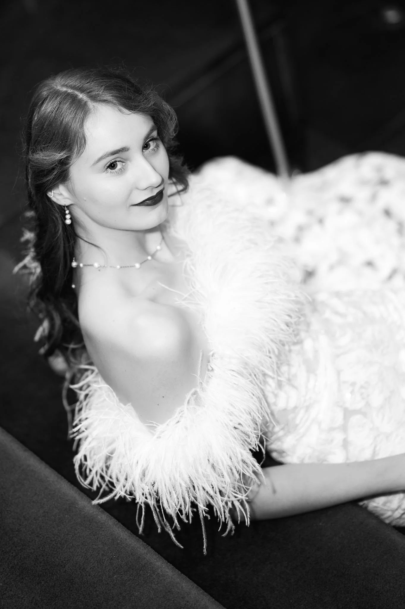 Junge Frau in eleganter Kleidung mit Federmantel, auf schwarzem Sofa, in Schwarz-Weiß-Fotografie.