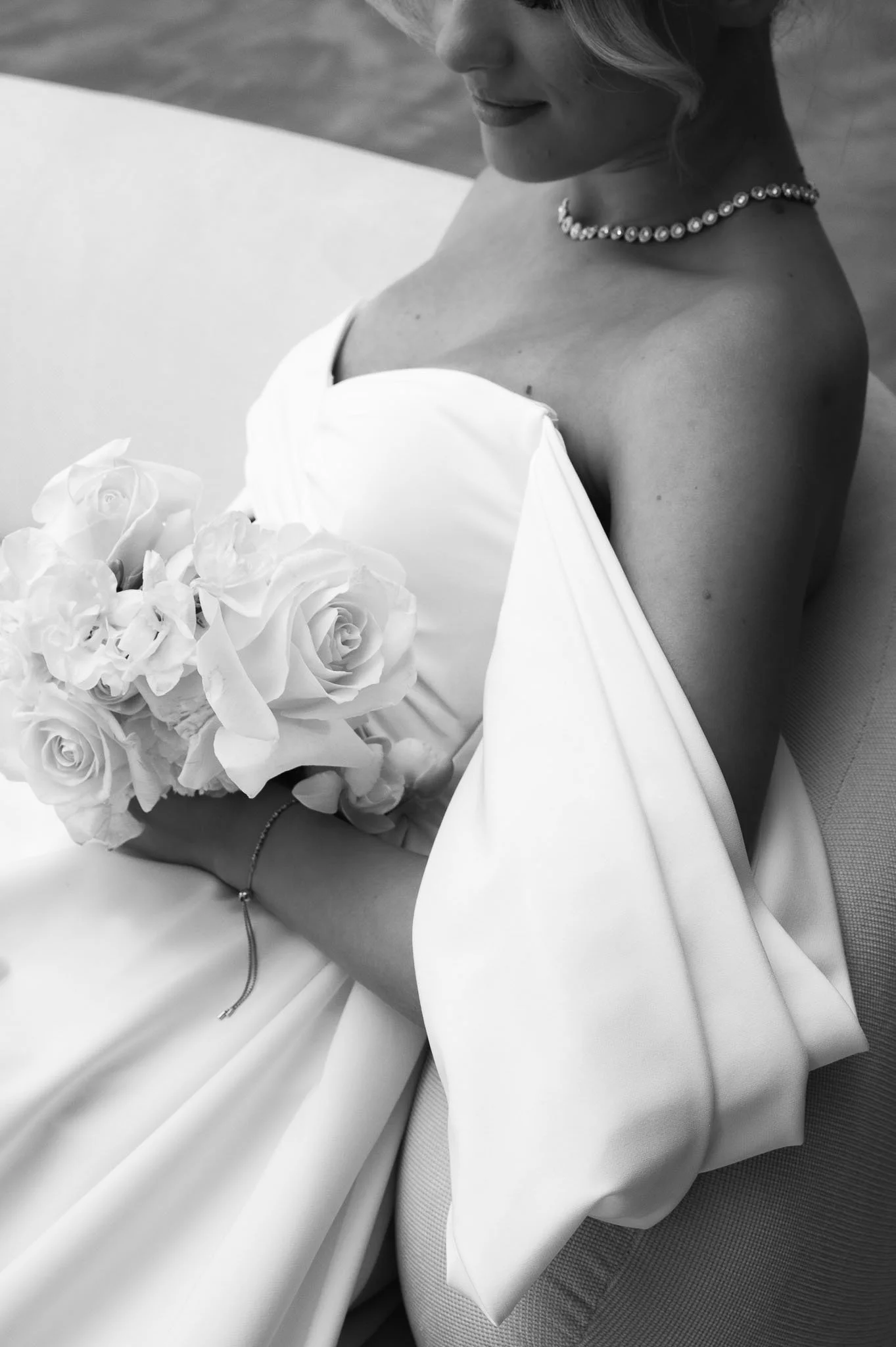 Naheaufnahme einer Braut in einem weißen Hochzeitskleid, die ein Bouquet mit Rosen hält, auf einem Stuhl sitzend. Die Braut trägt eine Perlenkette und ein Armband. Das Bild ist schwarz-weiß.