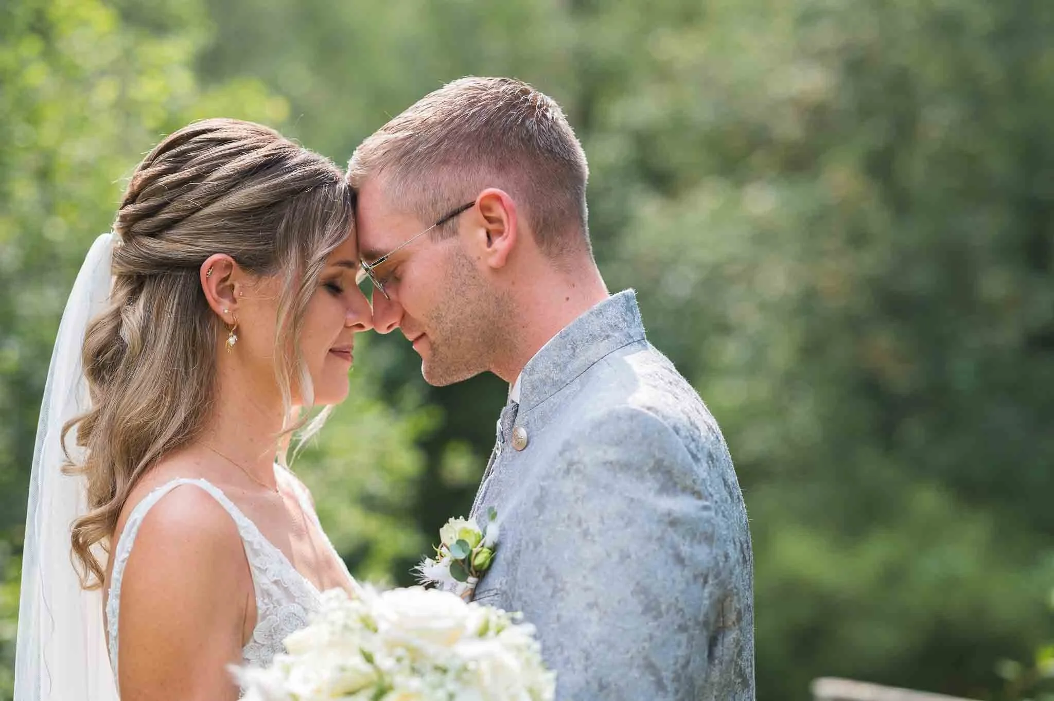 Braut und Bräutigam bei einer Hochzeit, die mit geschlossenen Augen und Stirn an Stirn in einer grünen, natürlichen Umgebung stehen.