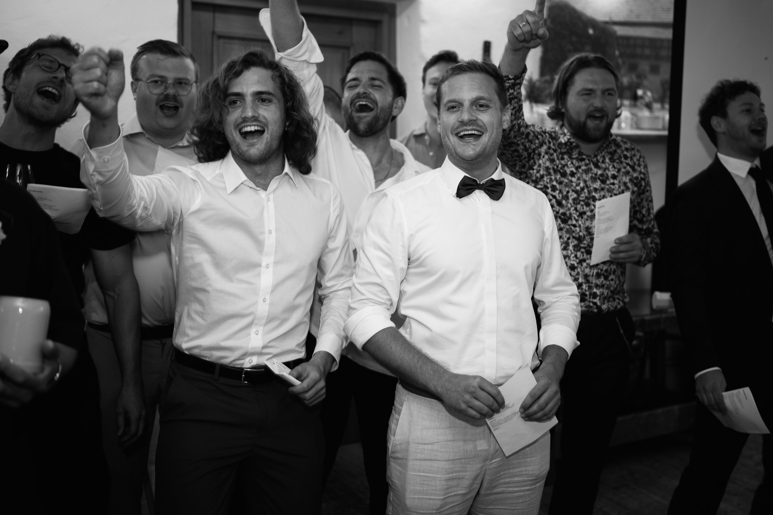 Group of people at a celebration or party, smiling and cheering, with some holding papers. One man in the foreground is wearing a tuxedo with a bow tie. The photo is in black and white.