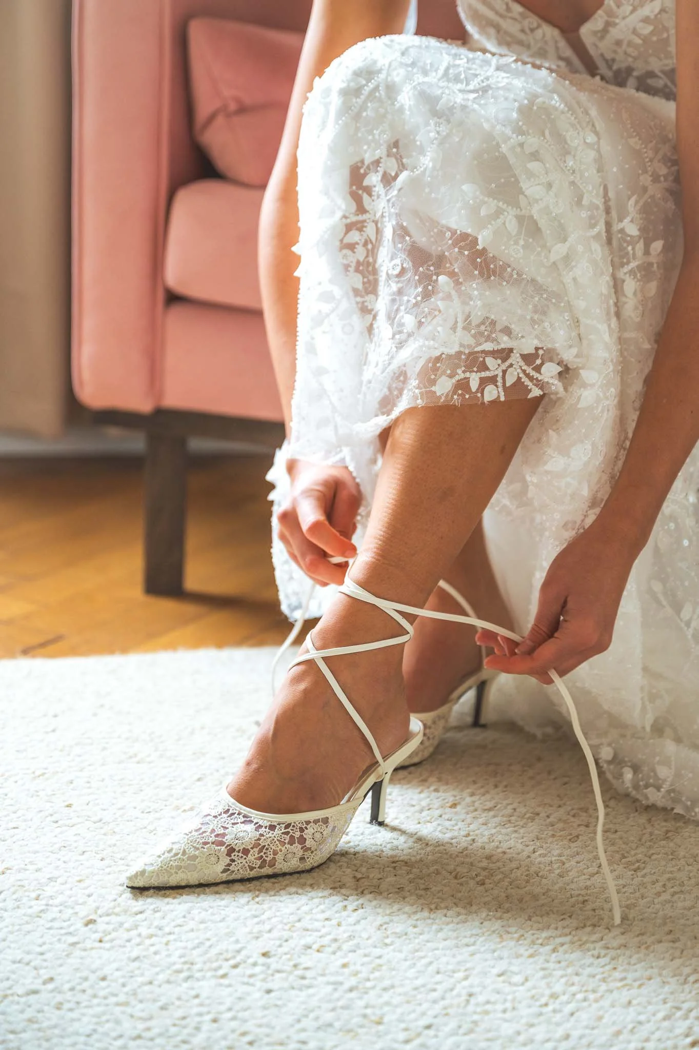 Eine Frau in einem weißen Brautkleid bindet die Schnürung ihrer weißen Spitzenschuhe an ihrem Knöchel, im Hintergrund ein pinkes Sofa.