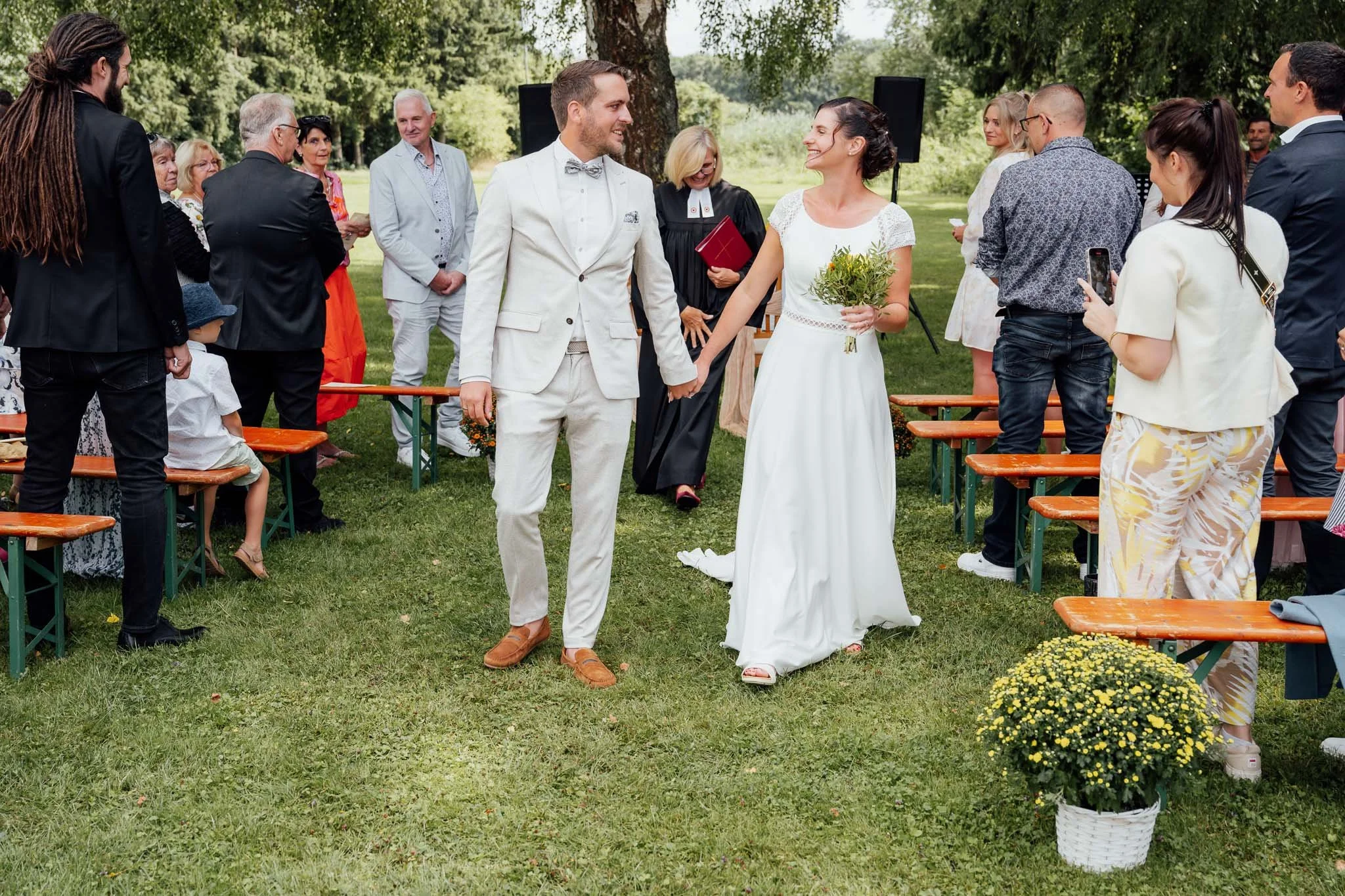 Ein Hochzeitsdinner im Freien mit lachendem Brautpaar, das sich die Hand hält, umgeben von Gästen auf grüner Wiese unter Bäumen.