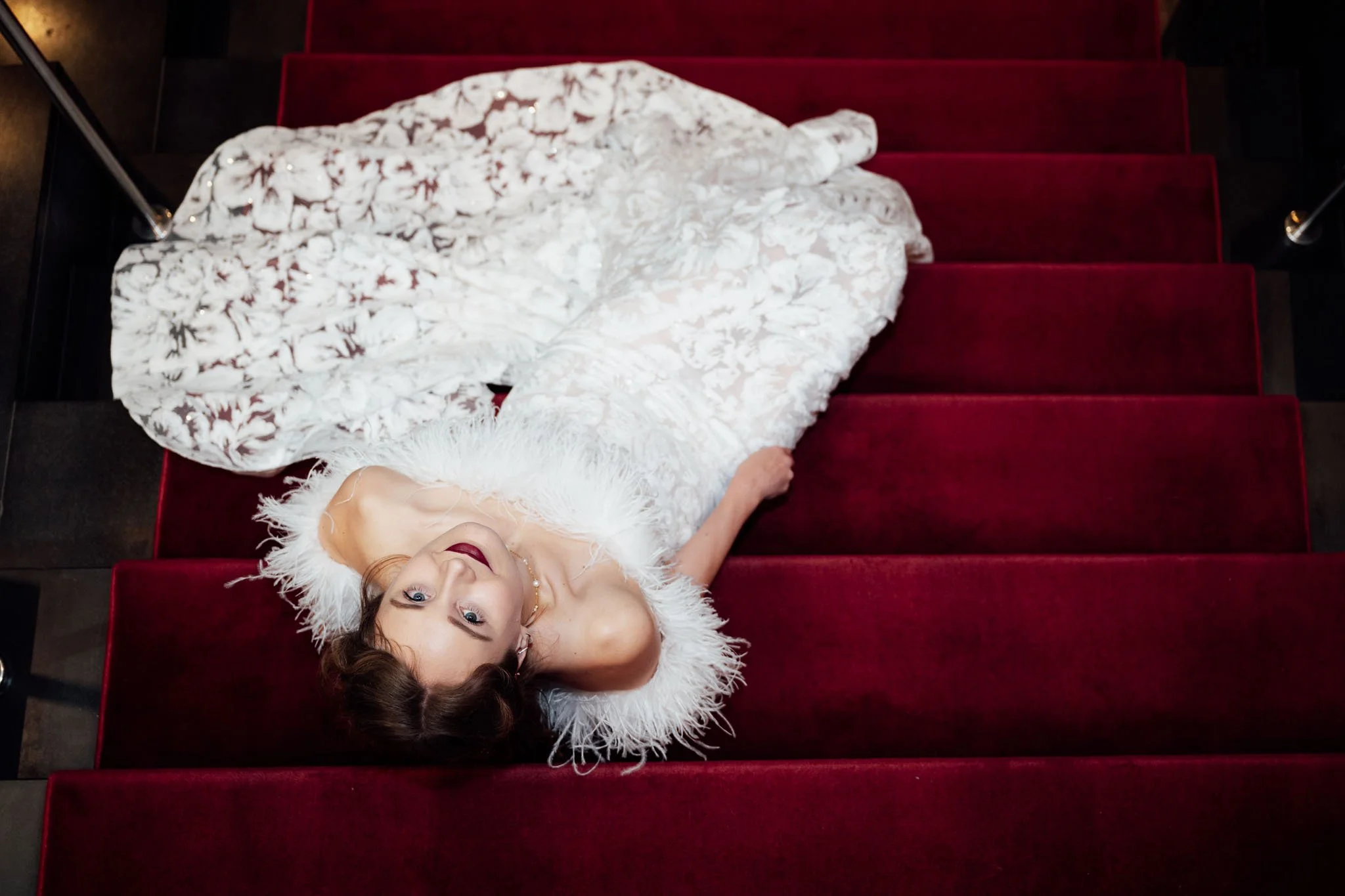 Frau in weißem, eleganten Kleid mit Federschmuck und rotem Lippenstift liegt auf roten Treppen und schaut in die Kamera.