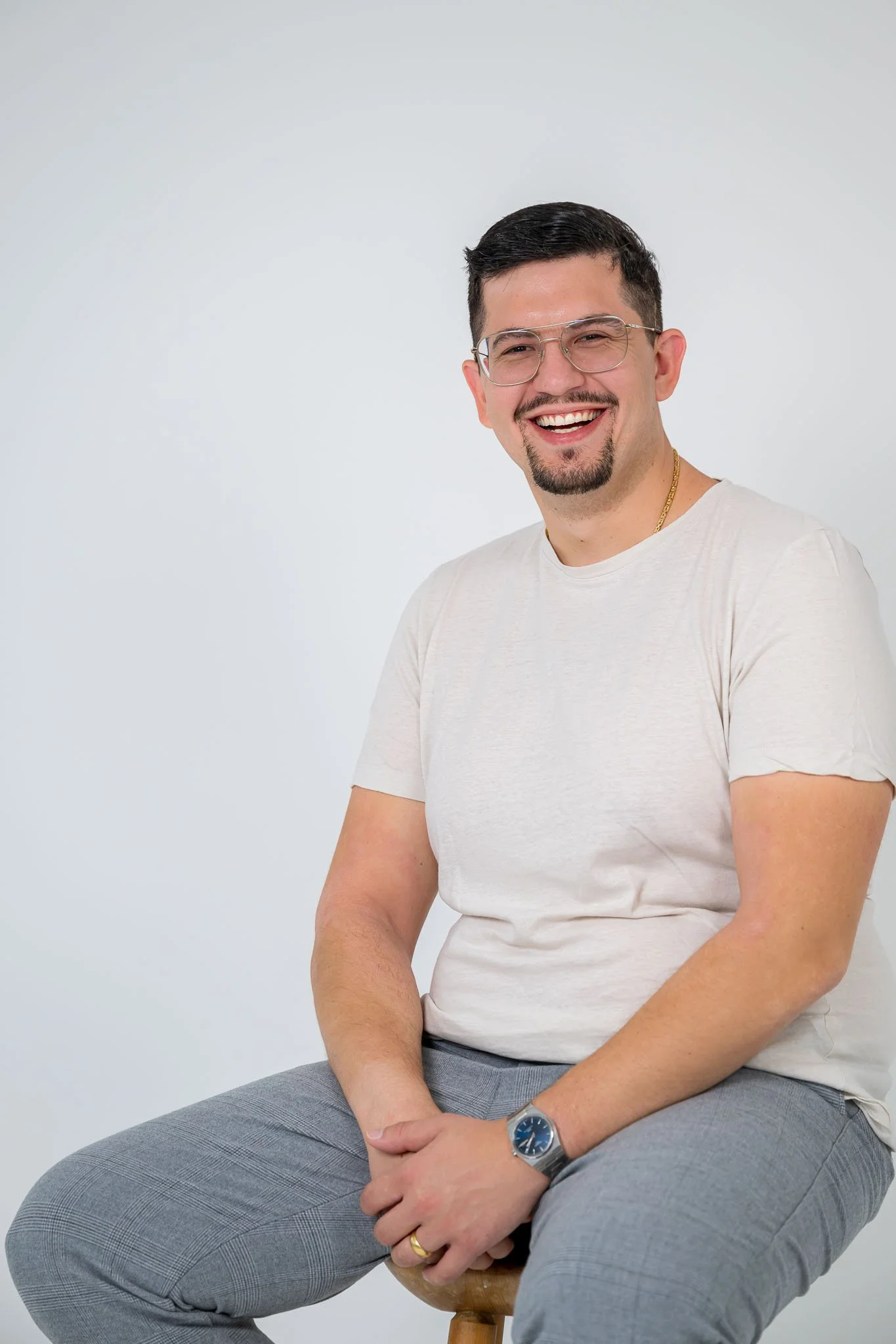 Der Fotograf Anto Lukic. Lächelnder Mann mit schwarzen Haaren, Brille, weißem T-Shirt, grauen Hosen, sitzt auf einem Hocker vor weißem Hintergrund.