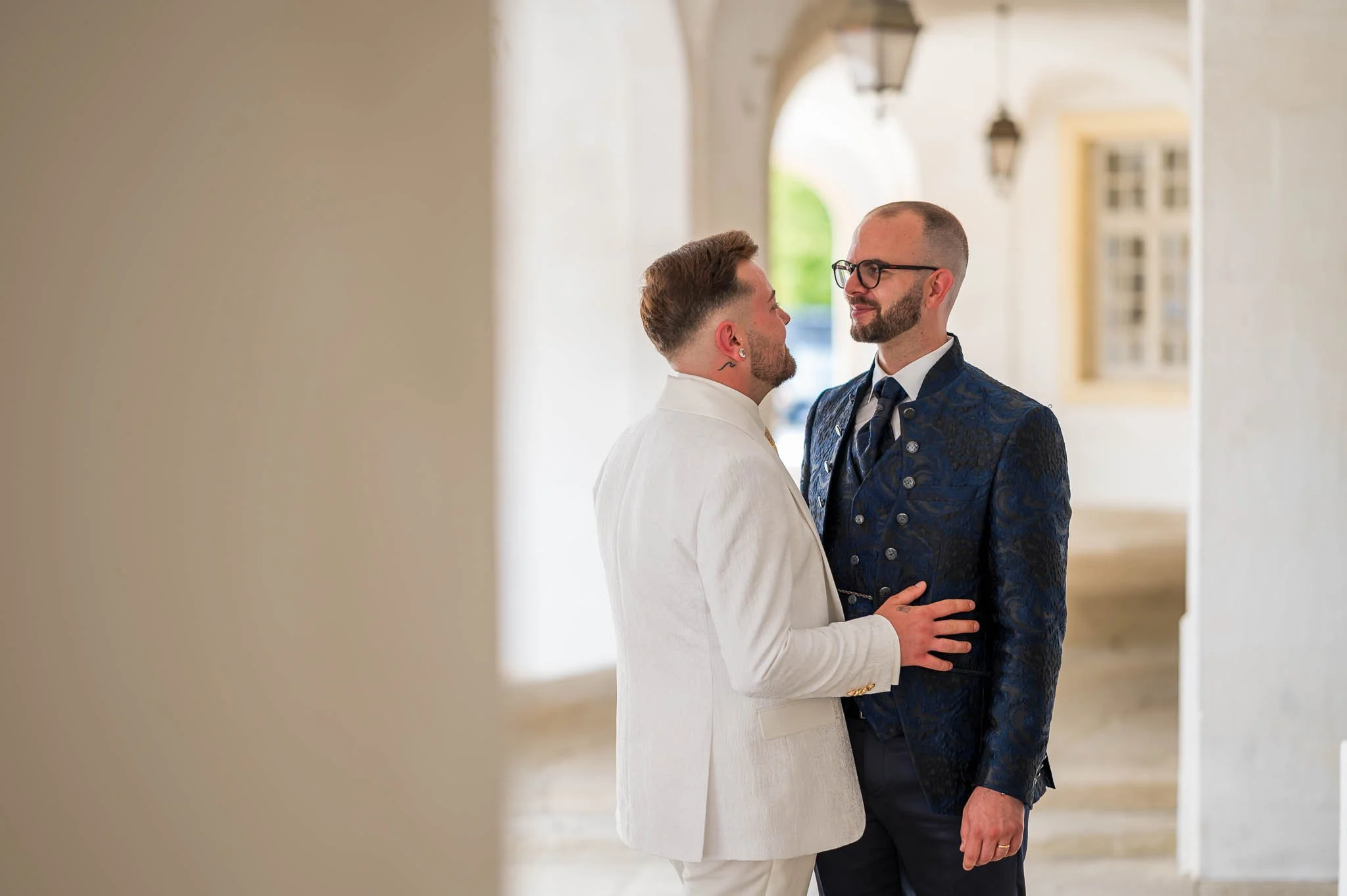 Zwei Männer, die sich bei einer formellen Gelegenheit in einem hellen, historischen Gebäude mit weißen Wänden und großen Fenstern gegenüberstehen, halten sich an den Händen und schauen einander an.