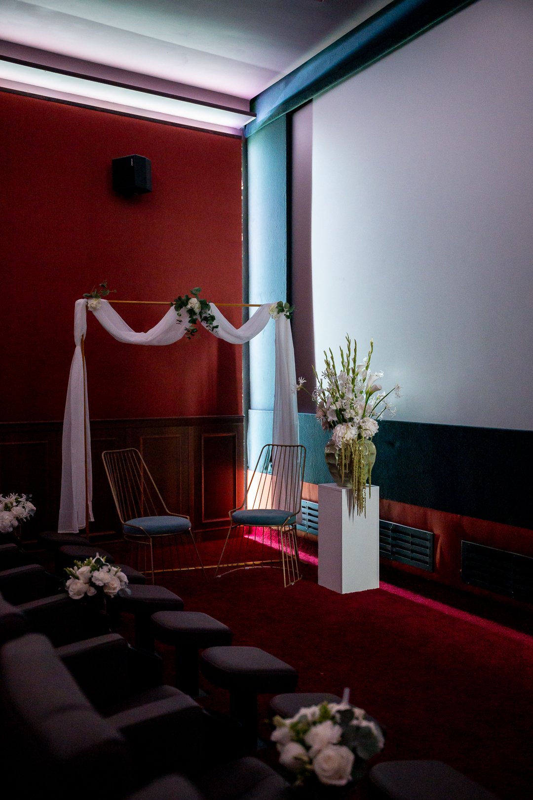 Ein Traubogen mit 2 Stühlen und Blumendeko vor der Leinwand in einem Kinosaal