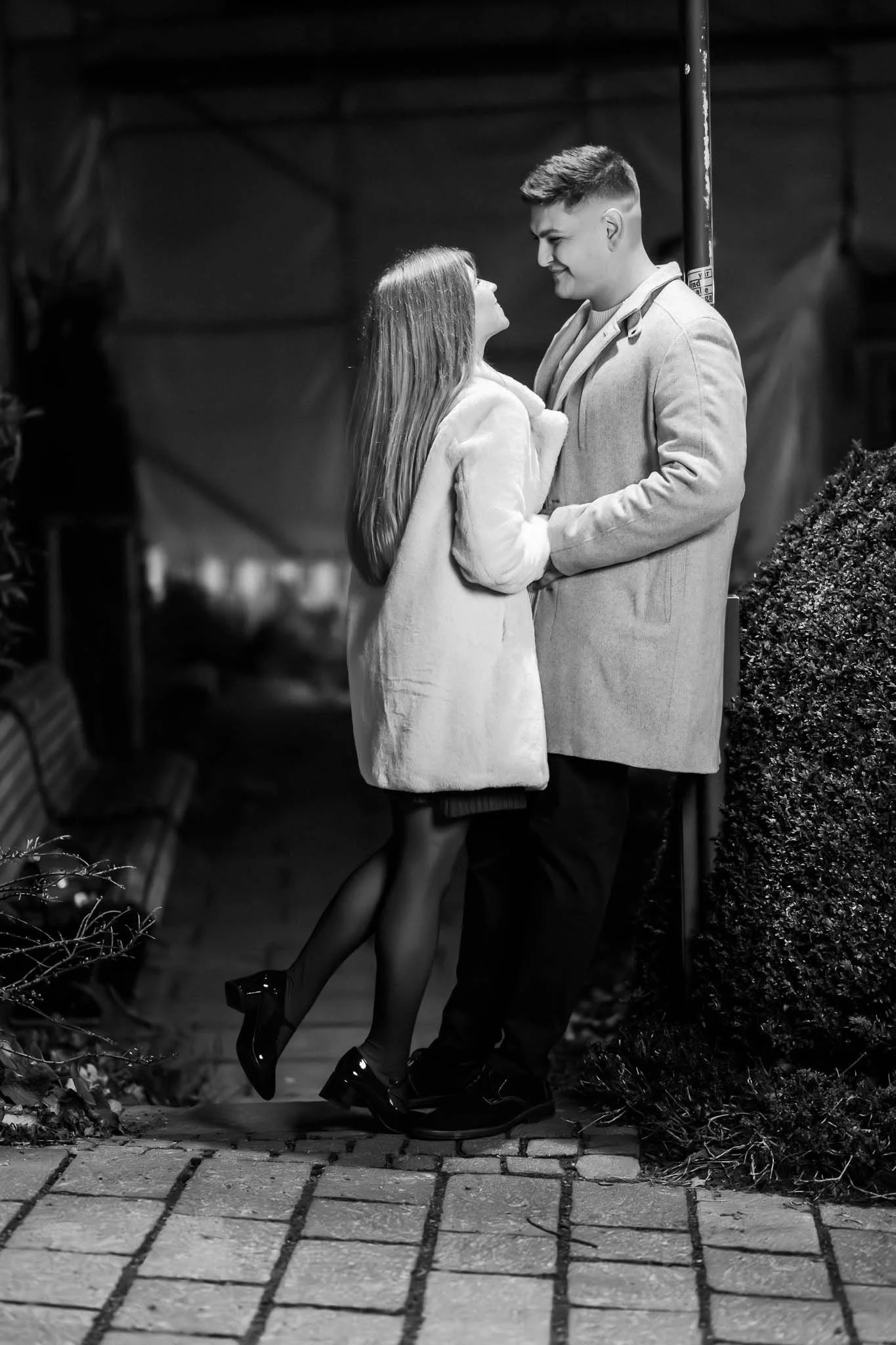 Schwarz-weiß Ganzkörperbild von einem Mann, der sich an einer Laterne anlehnt, während eine Frau auf Zehenspitzen versucht ihn zu küssen. Beide tragen Mäntel und lachen herzlich