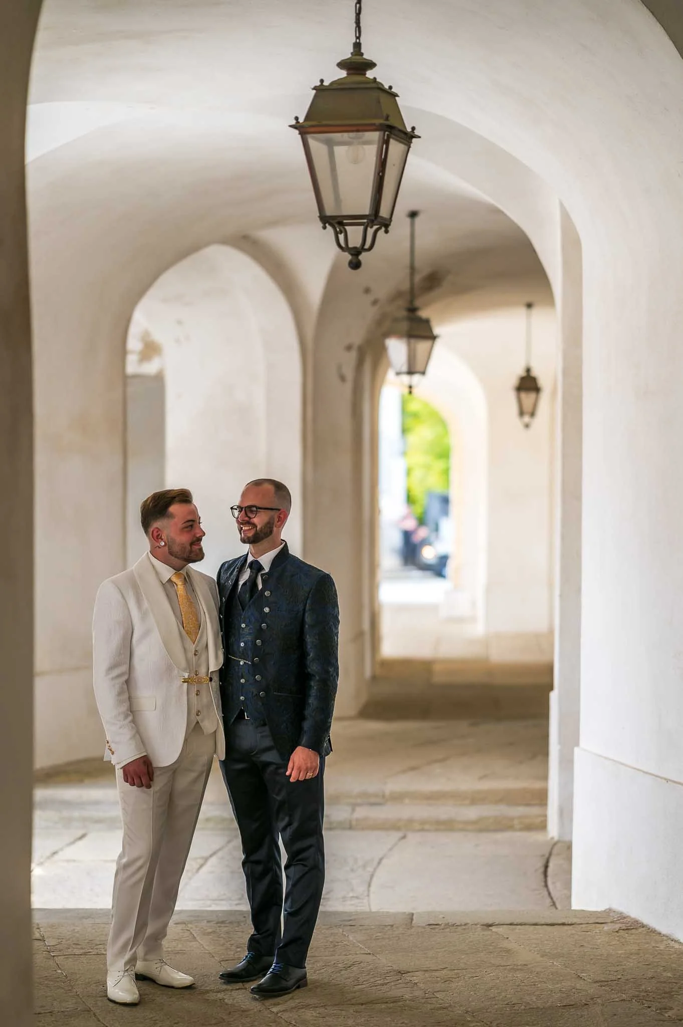 Zwei Männer in festlicher Kleidung stehen sich an einem eleganten Ort mit hohen, weißen Bögen und Laternen gegenüber, lächeln und schauen sich an.