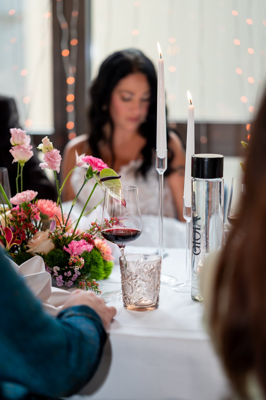 Nahaufnahme von einem Weinglas, welches aus einem festlich gedeckten Tisch steht bei Kerzenschein. Im Hintergrund ist eine Braut zu erkennen