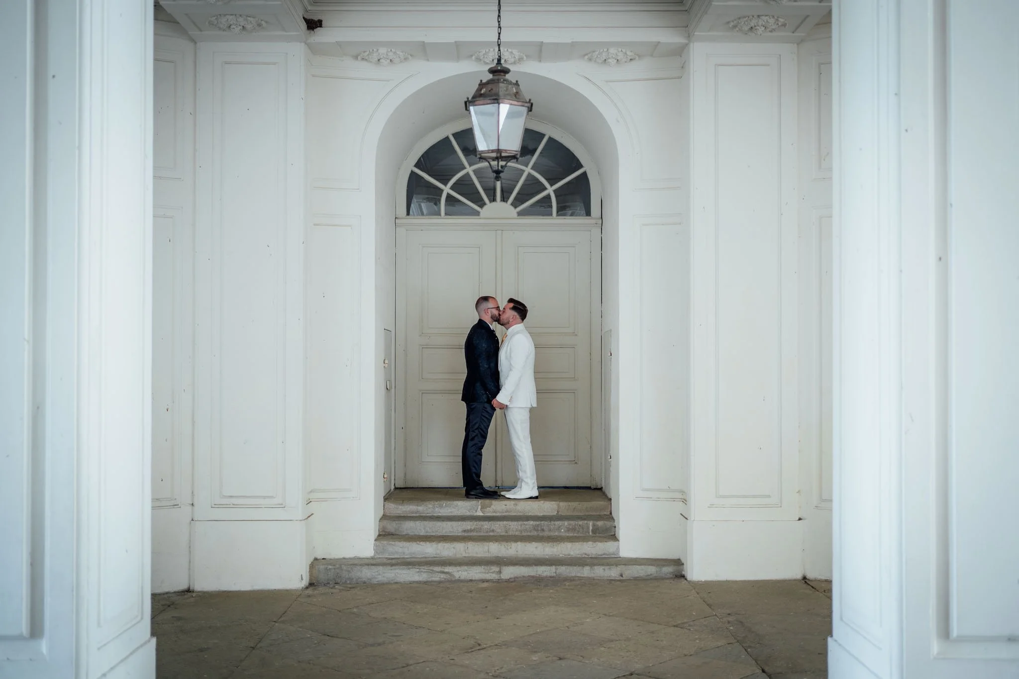 Zwei Männer in formellen Anzügen stehen sich auf den Stufen vor einer großen weißen Tür gegenüber, sie küssen sich.