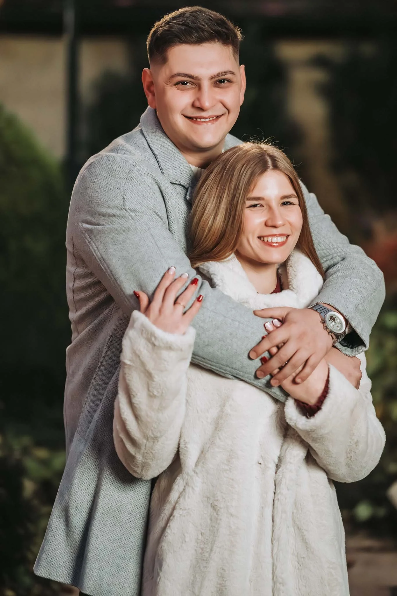 Porträt von einem Paar. Er steht hinter ihr und hat seine Hände über ihre Schultern gelegt. Beide blicken lachend in die Kamera und sie tragen Mäntel.