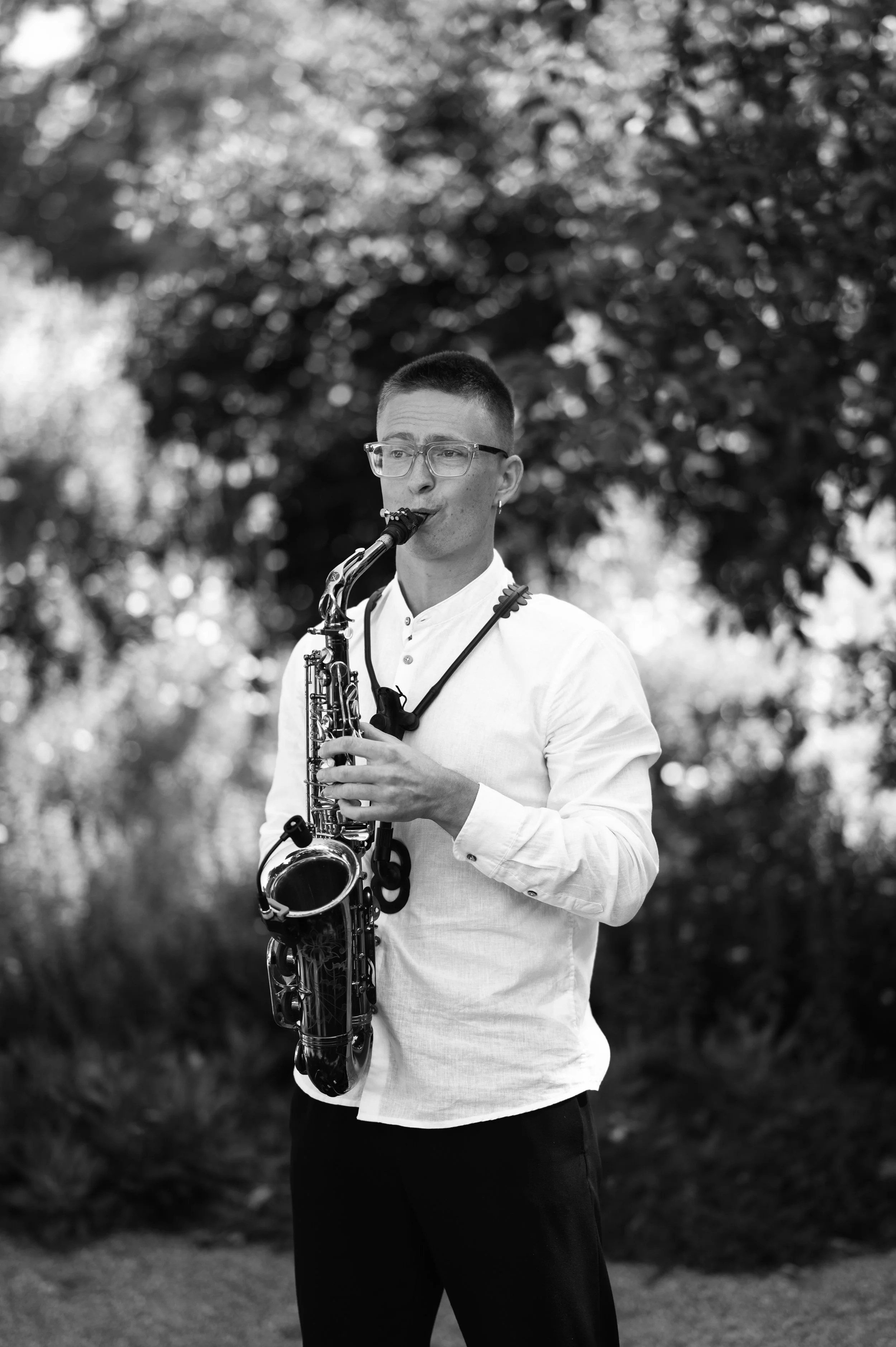 A young man with short hair and glasses playing a saxophone outdoors, dressed in a white shirt and black pants, with a blurred background of trees and foliage.