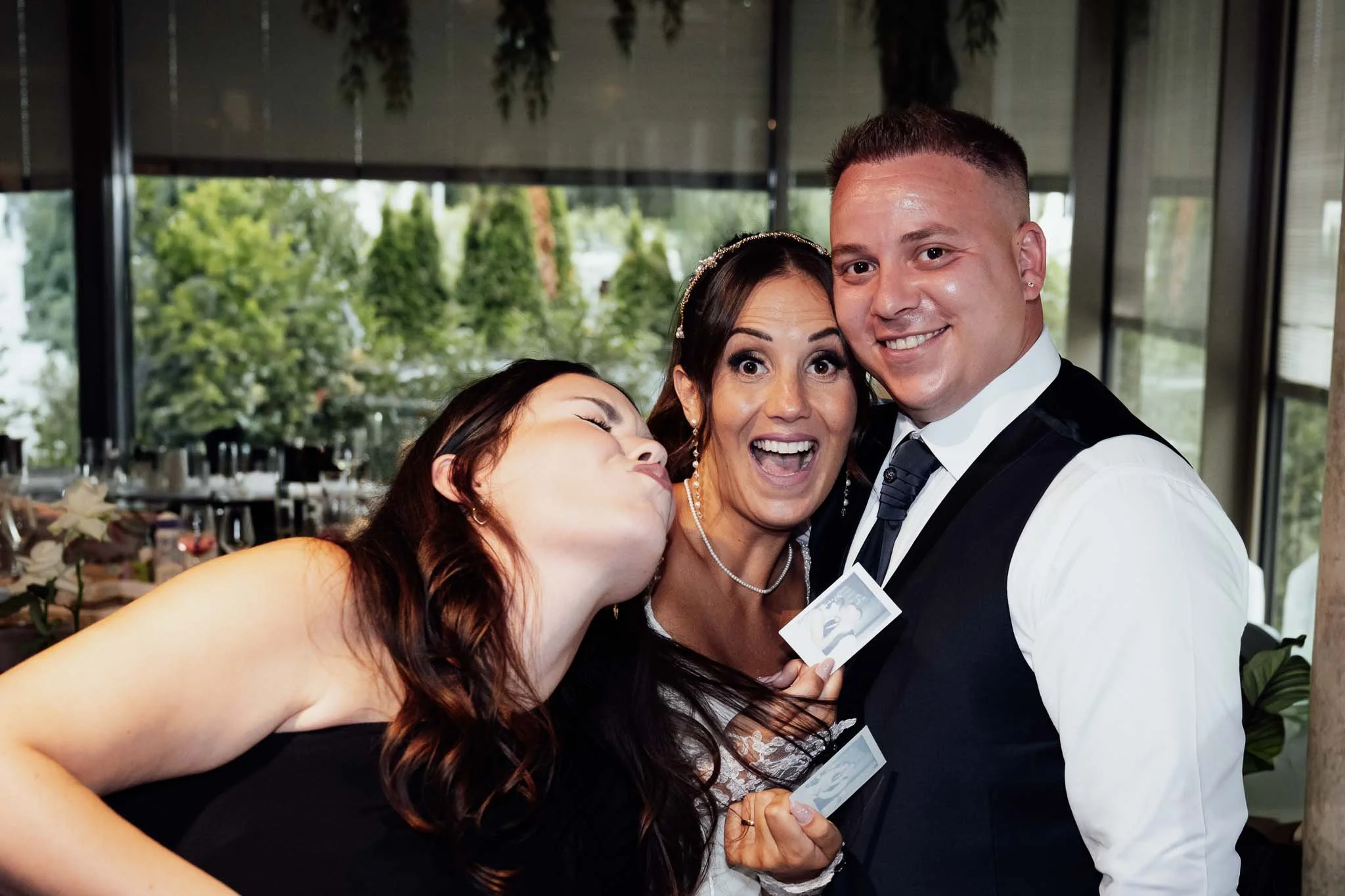 Ein Hochzeitsfest mit drei Personen, die freudig in die Kamera lächeln, inklusive einem Brautpaar und einem Freund, alle in formeller Kleidung, im Hintergrund eine moderne Veranstaltungsraum mit großen Fenstern und Grünpflanzen.