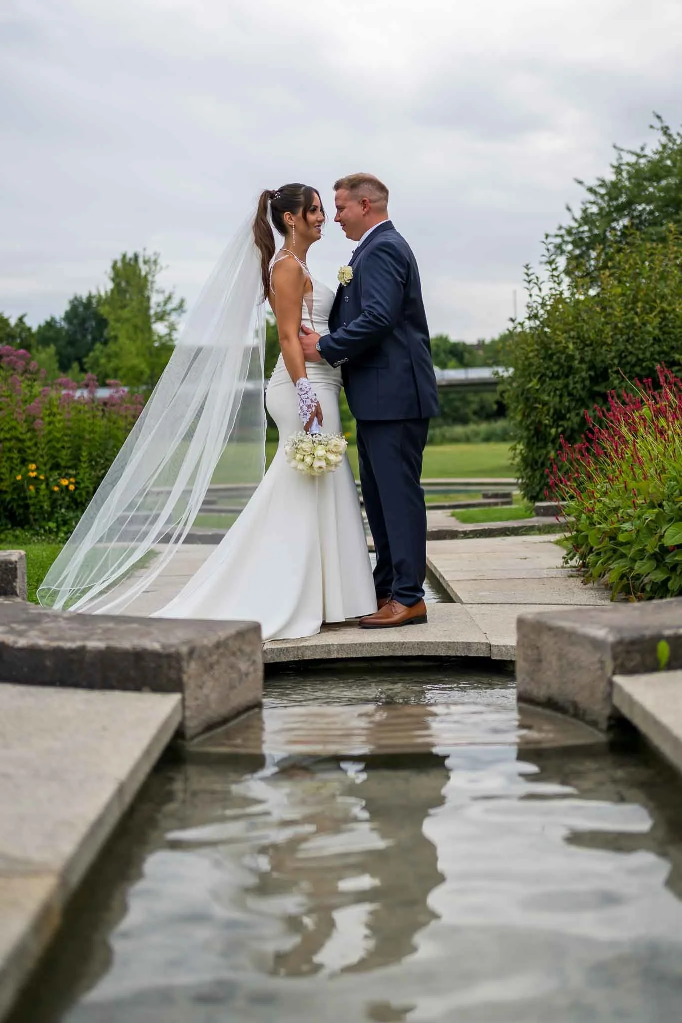 Ein Brautpaar steht sich auf einer kleinen Brücke über Wasser gegenüber, im Hintergrund ein Park mit Bäumen und Blumen, bei bewölktem Himmel.
