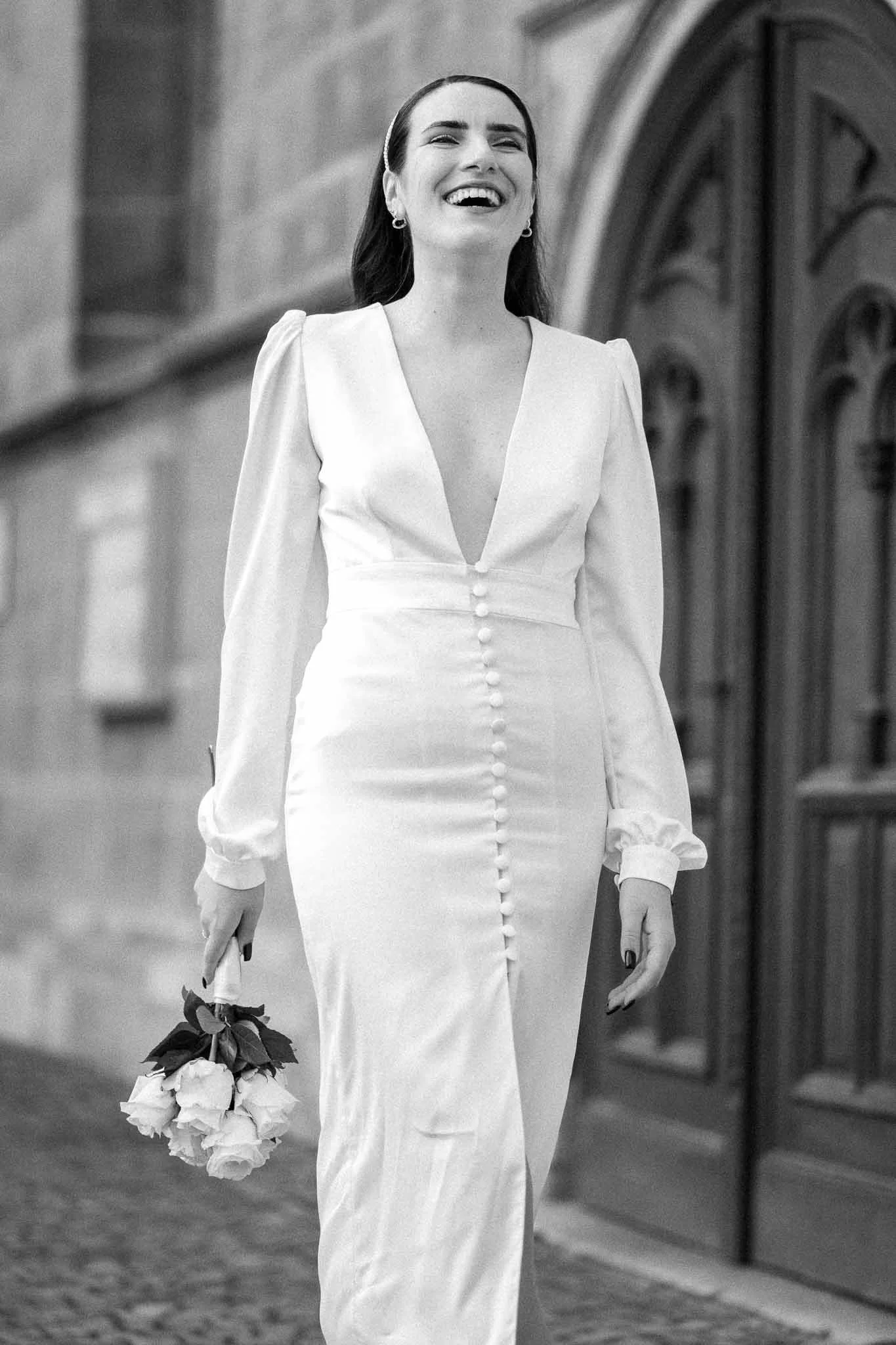 Schwarzweiß Bild einer Braut, die den Kopf in den Nacken gelegt hat und herzlich lacht, während sie läuft. Dabei hält sie einen kleinen Blumenstrauß aus Rosen in der Hand